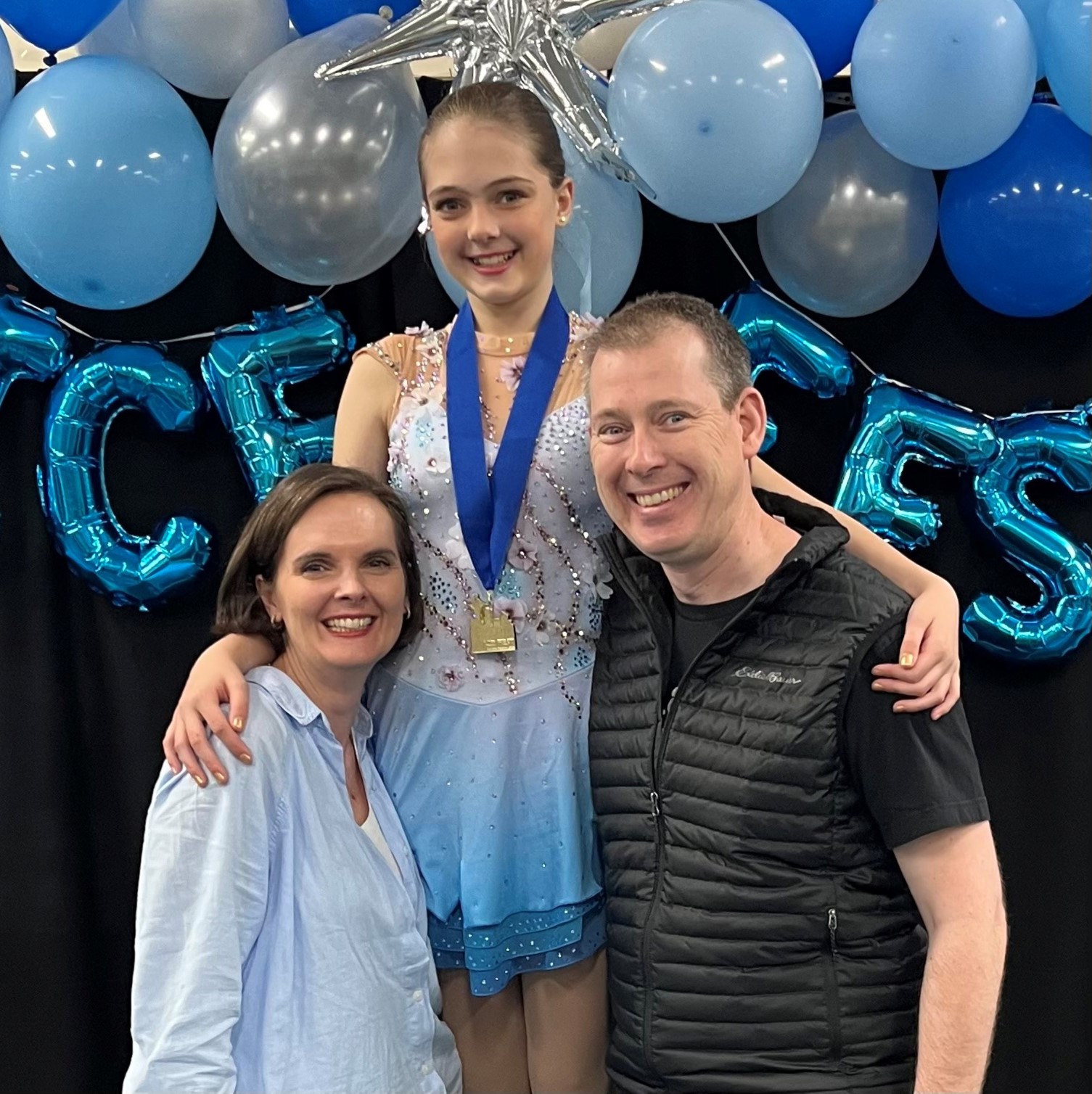 Odessa Akervall, dressed in her white and blue skating dress and with a medal around her neck, stands on the podium with her parents.