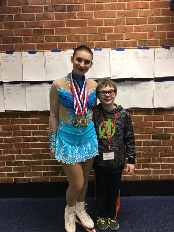 Marcus and his older sister at her high school competition in 2018. His sister (left) stands with her arm around Marcus. She has her hair slicked back in a bun and she is wearing a bright blue skating costume. Marcus (right) is a young boy with glasses wearing an Avengers hoodie and sweat pants