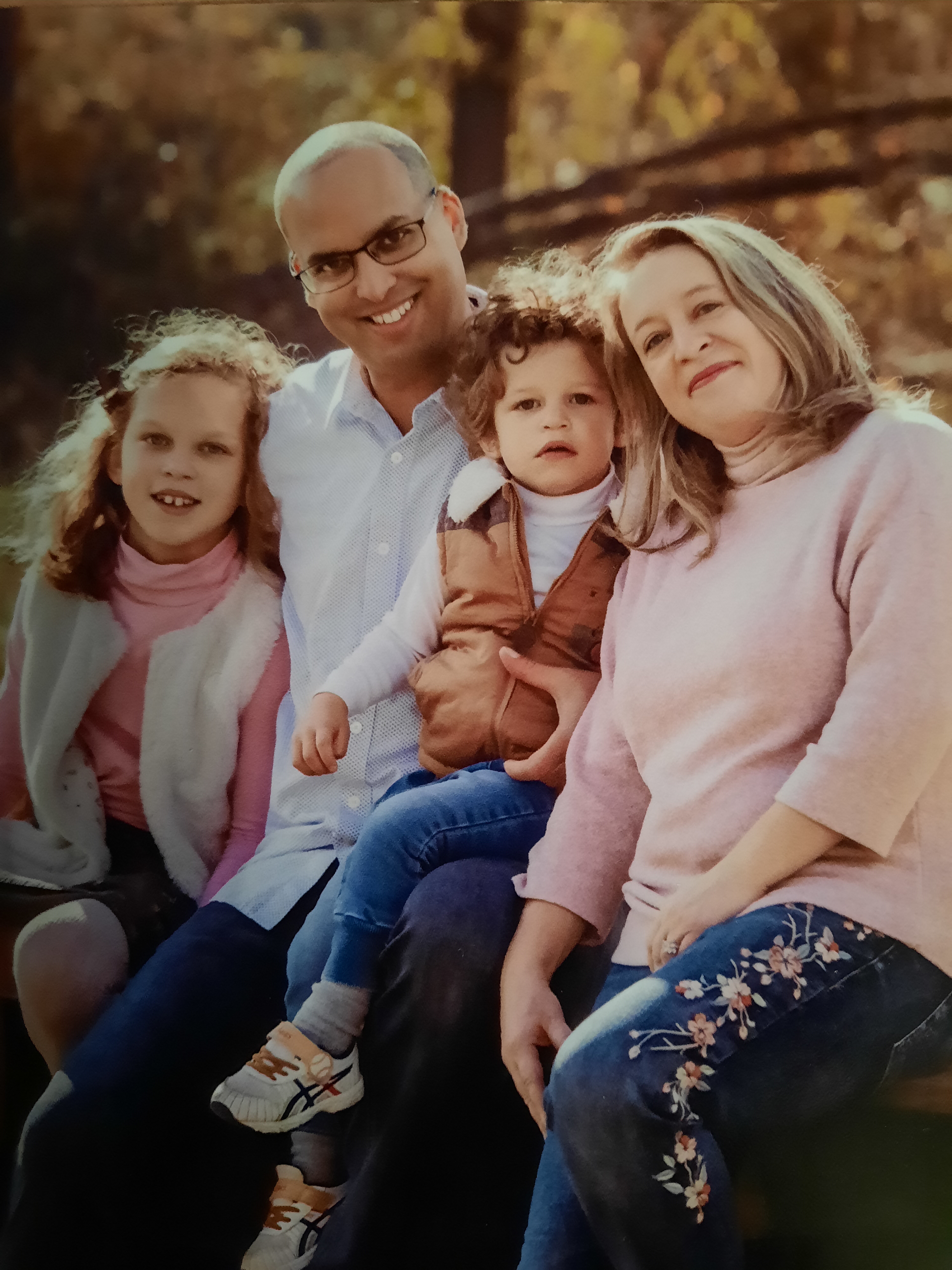 Aaron Parchem poses for a photo with his eife and their young daughter and son. Parchem is wearing a white button down shrit and glasses. His wife who stands on the far right side of the photo is wearing a pink turtleneck and jeans embroidered with flowers. Their won sittng between them has brown curly hair and is wearing a white long sleeve shirt, brown vest and jeans. Their daughter who sits to the left of Aaron has shoulder length light brown hair and is wearing a pink long sleeve shirt, white vest and jeans