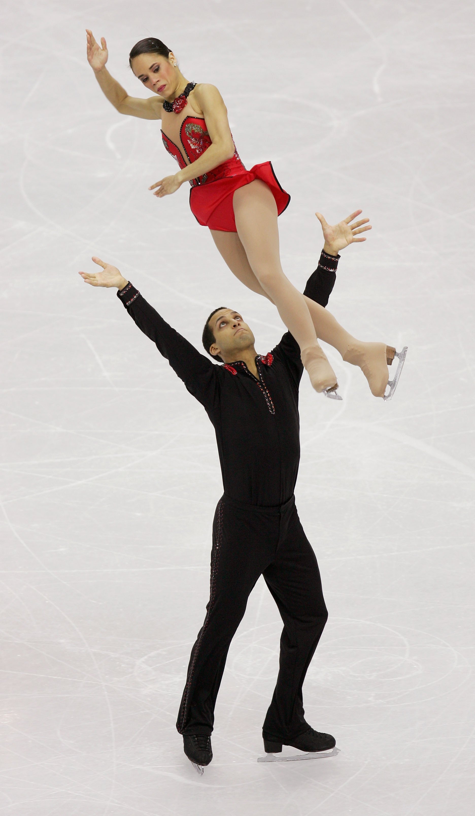 Aaron Parchem throws his partner Marcy Hinzmann in the air during a performance. Parchem is wearing a black long sleeve shirt with a red collar and black pants. Hinzmann is wearing a red skating costume with black trip along the neckline