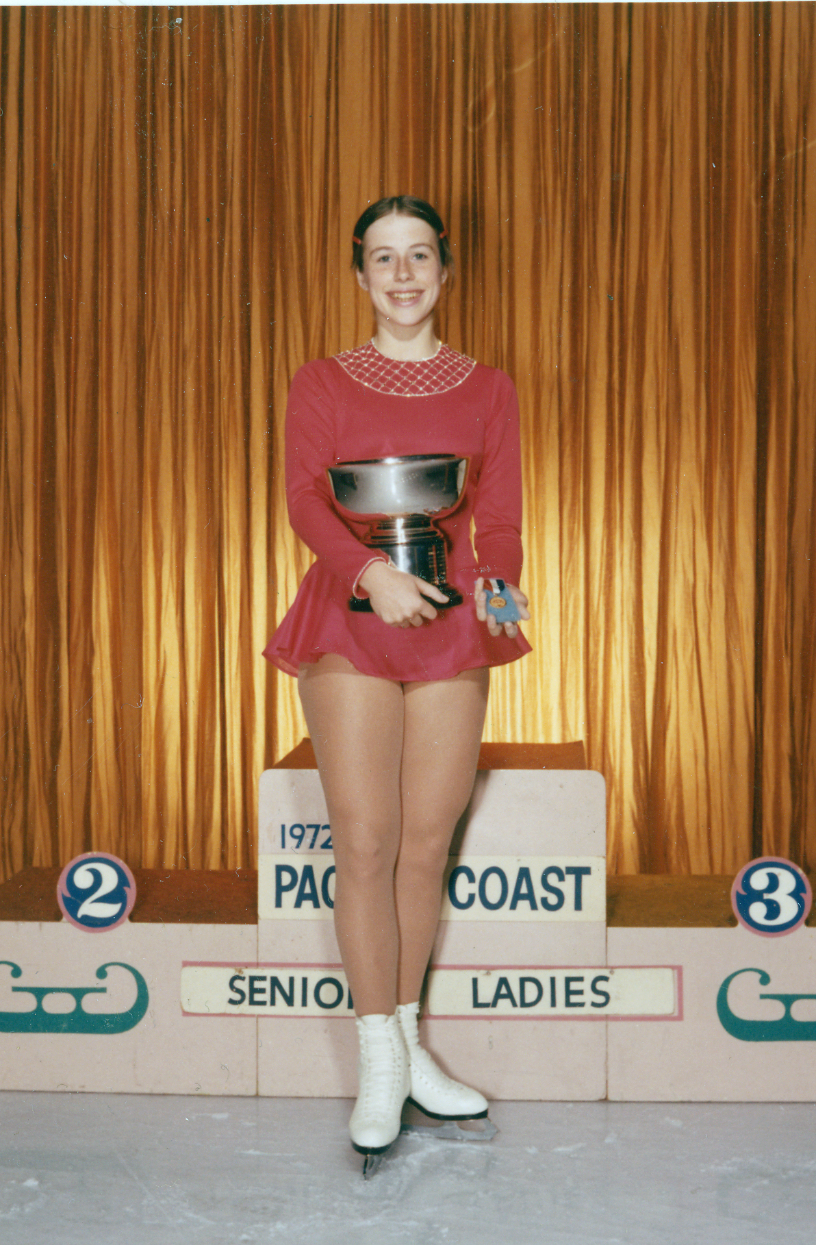 Patricia Shelley holds up a trophy on the podium post competition in 1972 wearing a faded red dress.