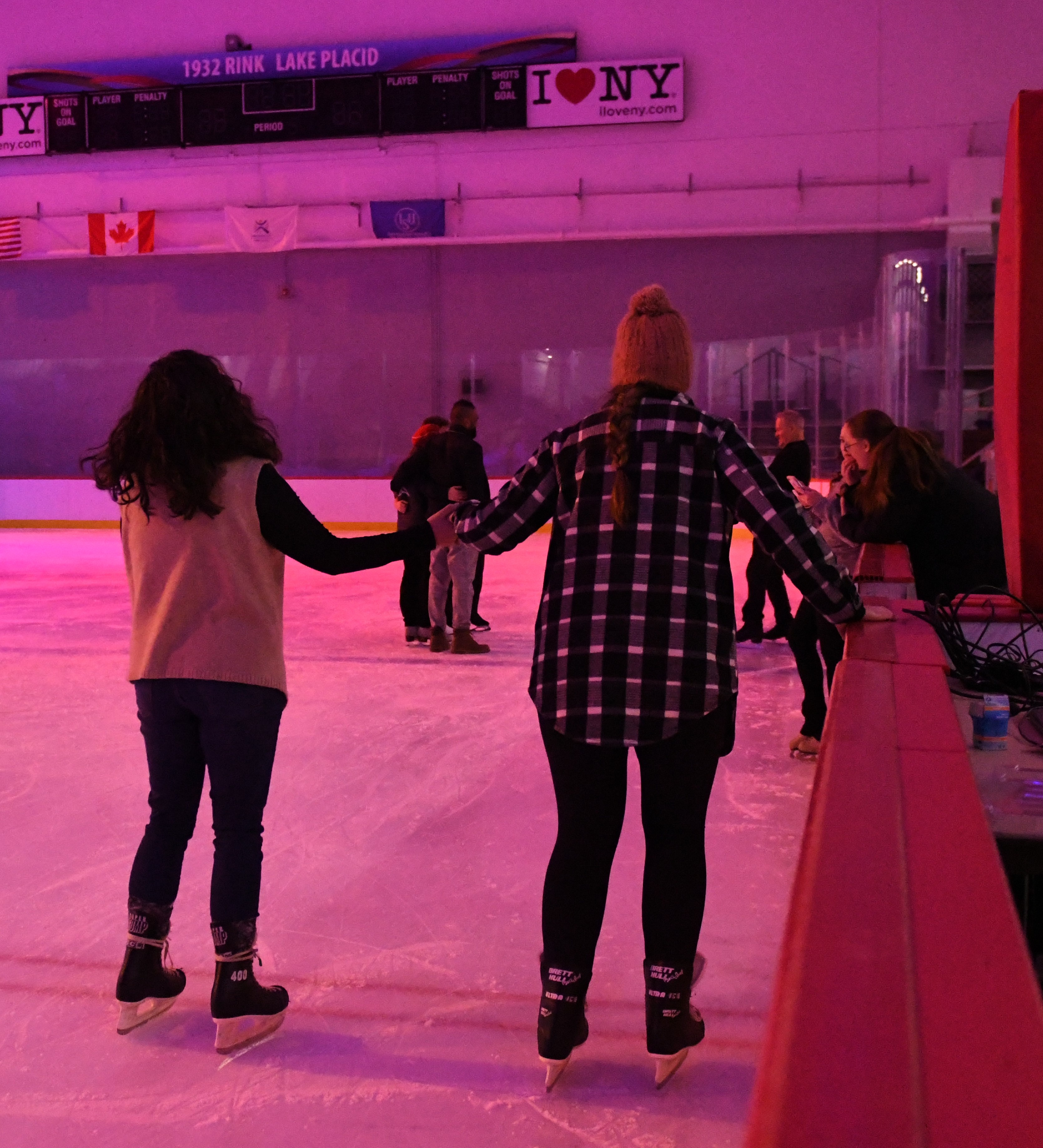 Photo taken from behind of two skaters on the ice holding hands. One is wearing a plaid shirt and yellow beanie while the other is wearing a white vest over a black long sleeve shirt