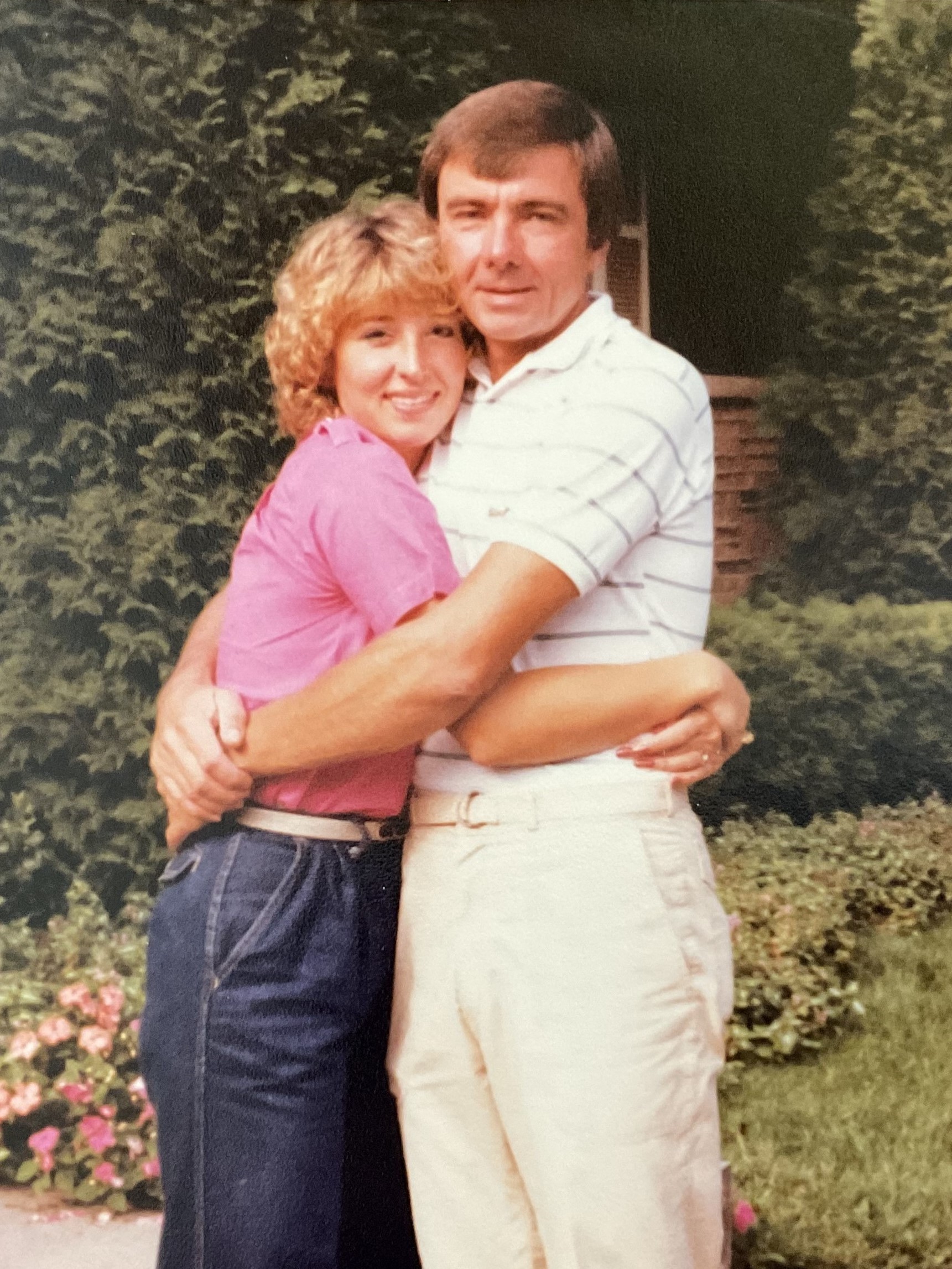 Sandy hugs her father in a photo taken in the late 1970s. Sandy (left) has blonde curly hair and is wearing a pink tshirt and jeans. Her father has on a white polo shirt and khakis. They both have their arms around each other