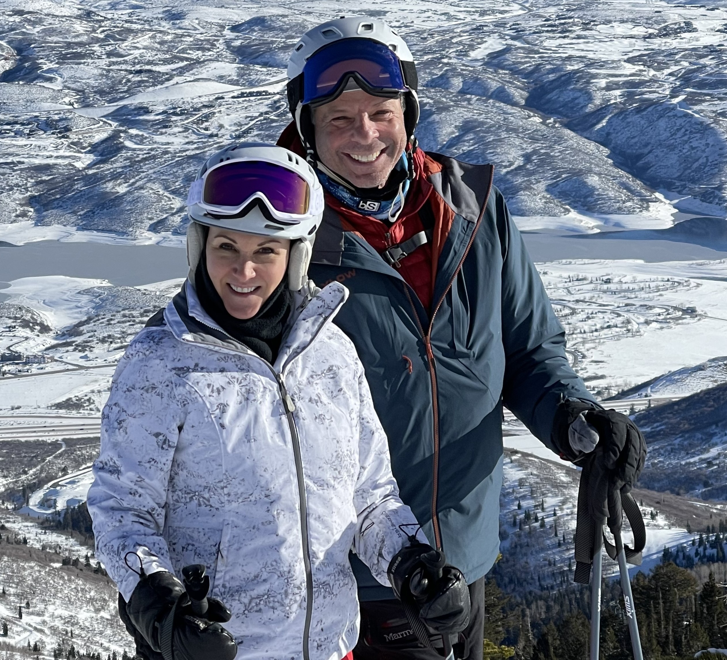 April Sargent Silverstein poses with her husband Larry while skiing. April is wearing a white ski jacket and has on a white helmet with goggles on the front. She holds two ski poles. Larry stands behind her wearing a blue ski jacket with a red jacket underneath. He wears a white helmet with goggles and holds ski poles. 