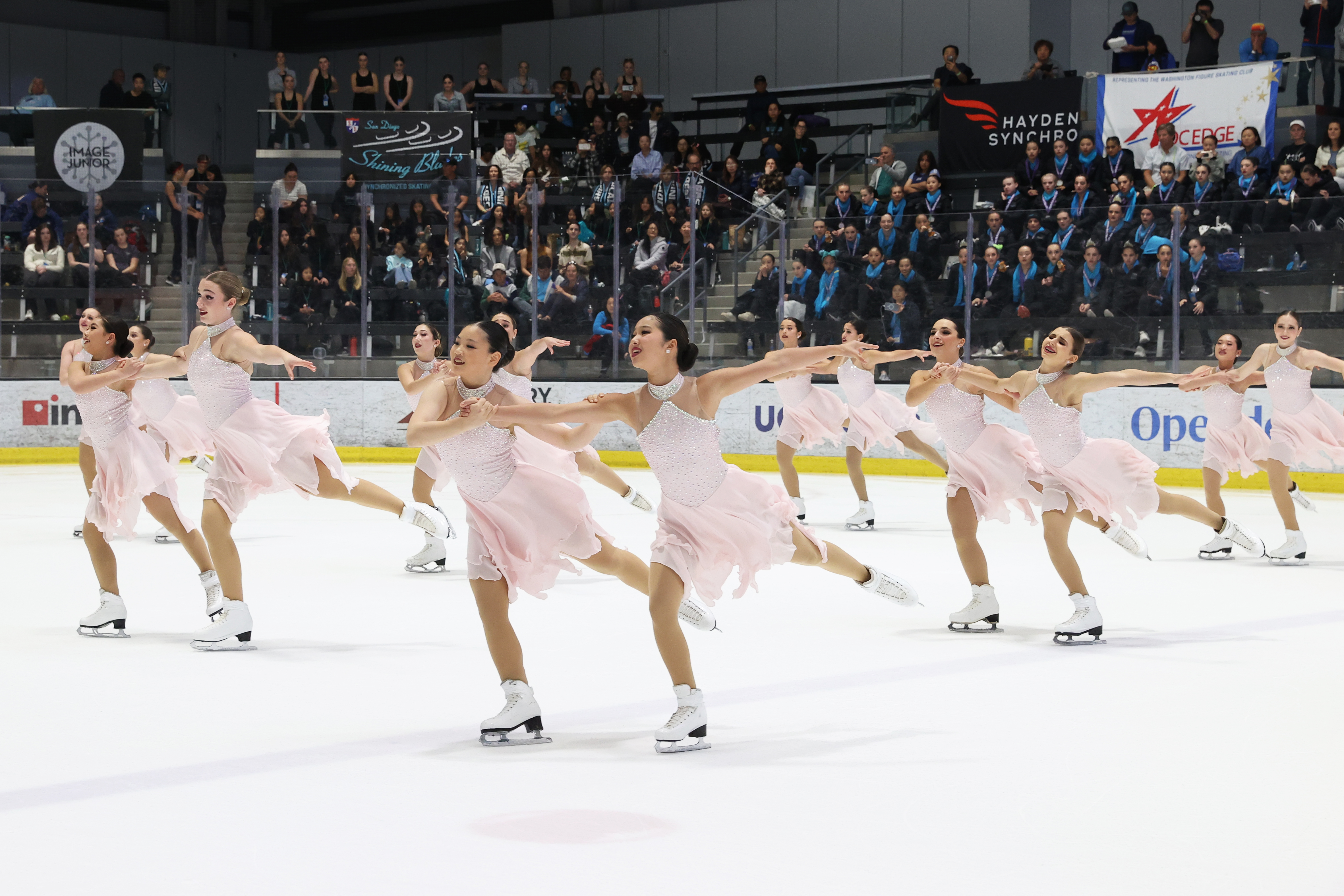 The Skyliners compete wearing matching light pink skating costumes. They all skate to the left with their arms outstretched and one leg extended behind them