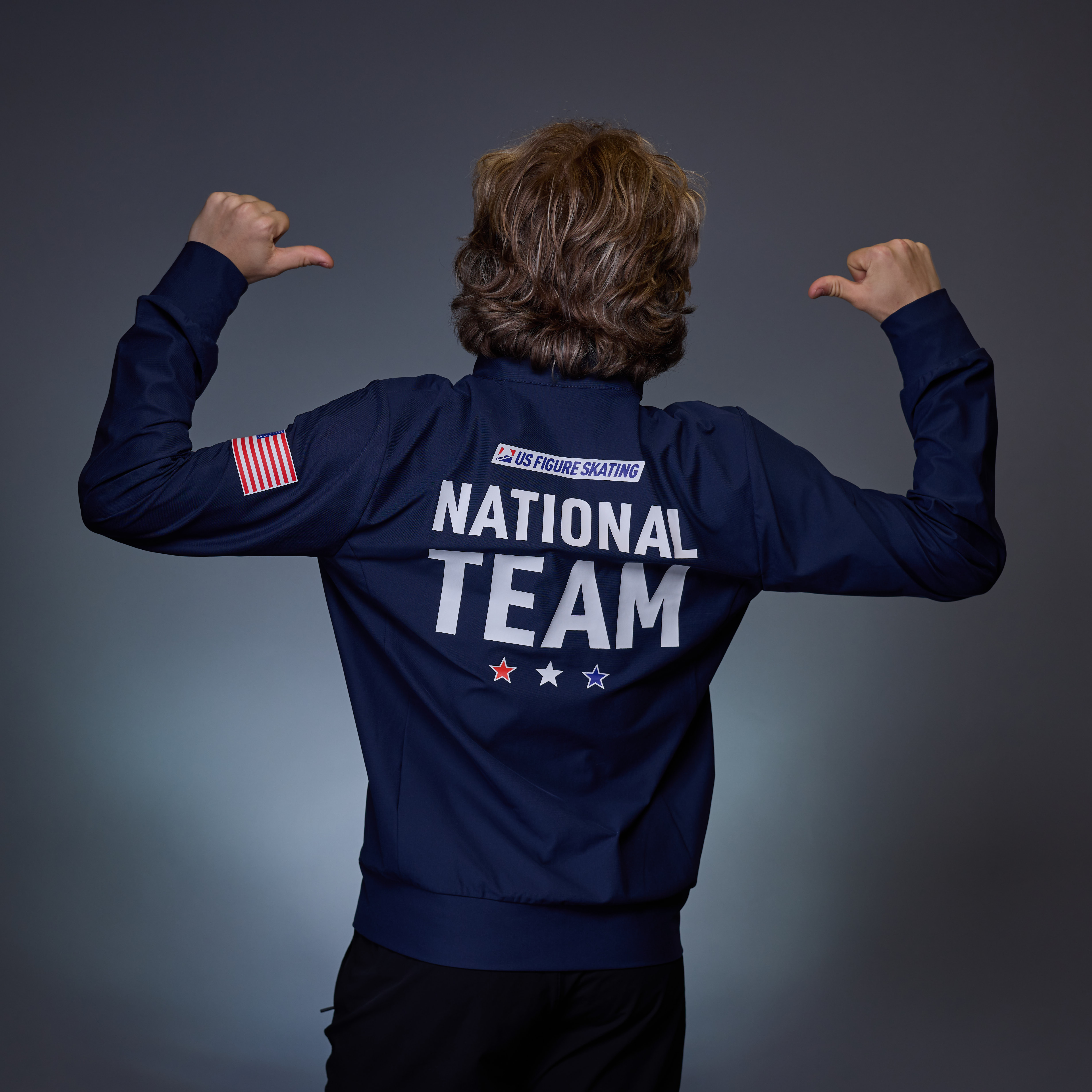 Ilia Malinin, with back turned, points back to the wording on his U.S. National Team jacket