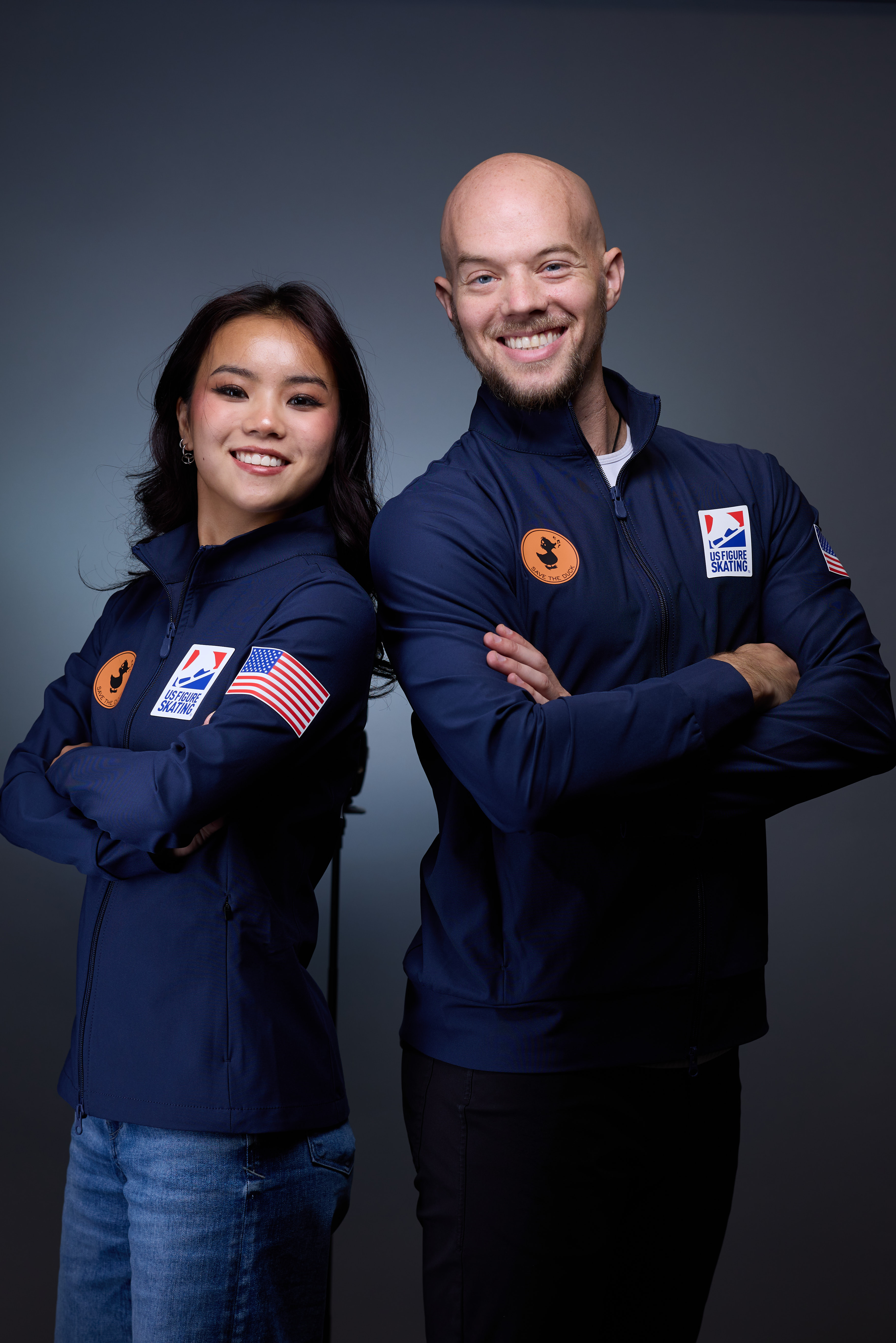 Ellie Kam and Danny O'Shea, wearing their blue U.S. National Team jackets, fold their arms and smile