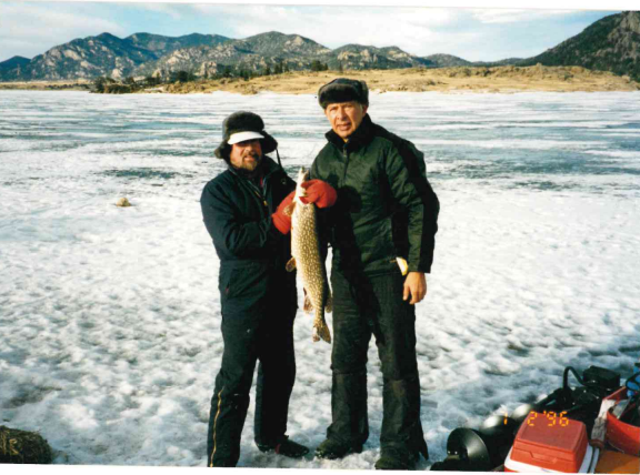 Photo of Bill holding a fish he caught. He is with  friend and they are both wearing black winter coats and black pants with fuzzy hats. 