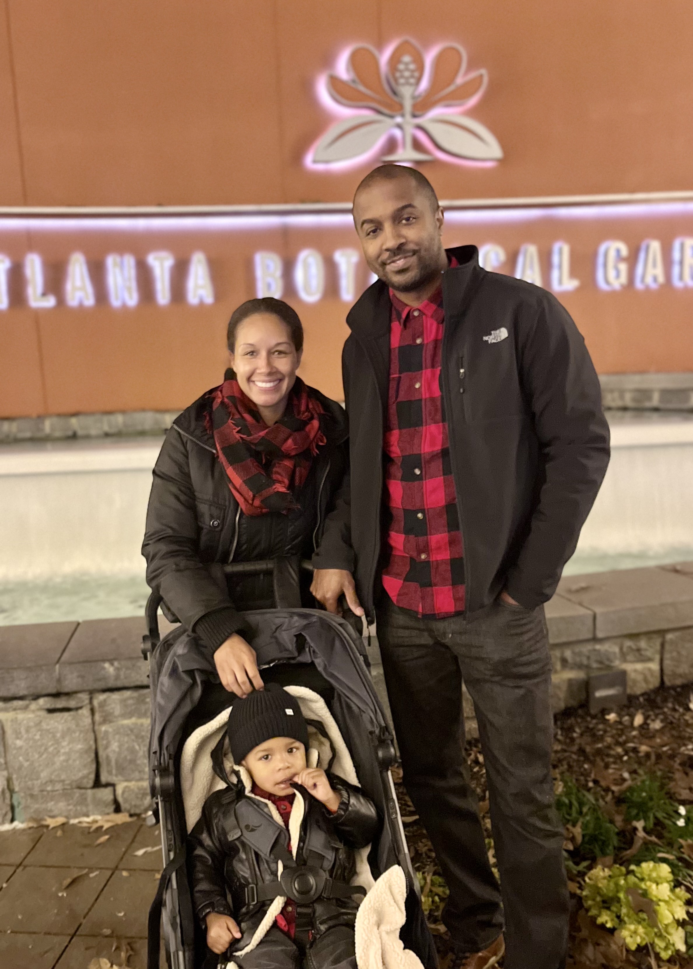 Cohen Duncan stands with her husband Darius and young son. She is a middle aged African American woman with hair tied back in a ponytail. She is wearing a black jacket with a red and black checkered scarf. Darius stands to her right wearing a red and black checkered shirt with a black jacket over the top. Their son sits in front of them in a stroller wearing a black beanie and leather jacket