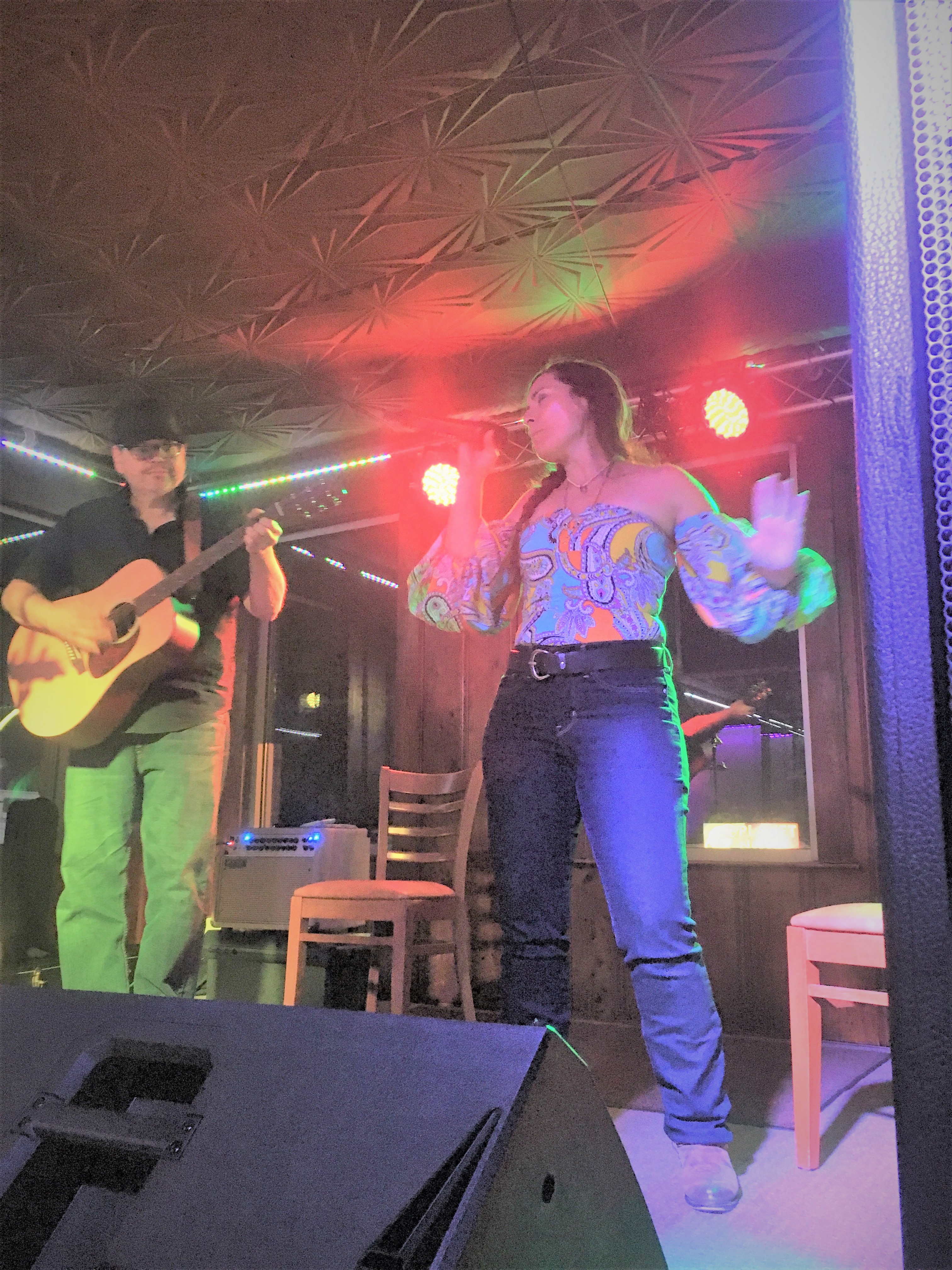 Suzie Flynn in a floral flowy top and a brown braid sings into a microphone while Stephen France sits on a bench playing the guitar.