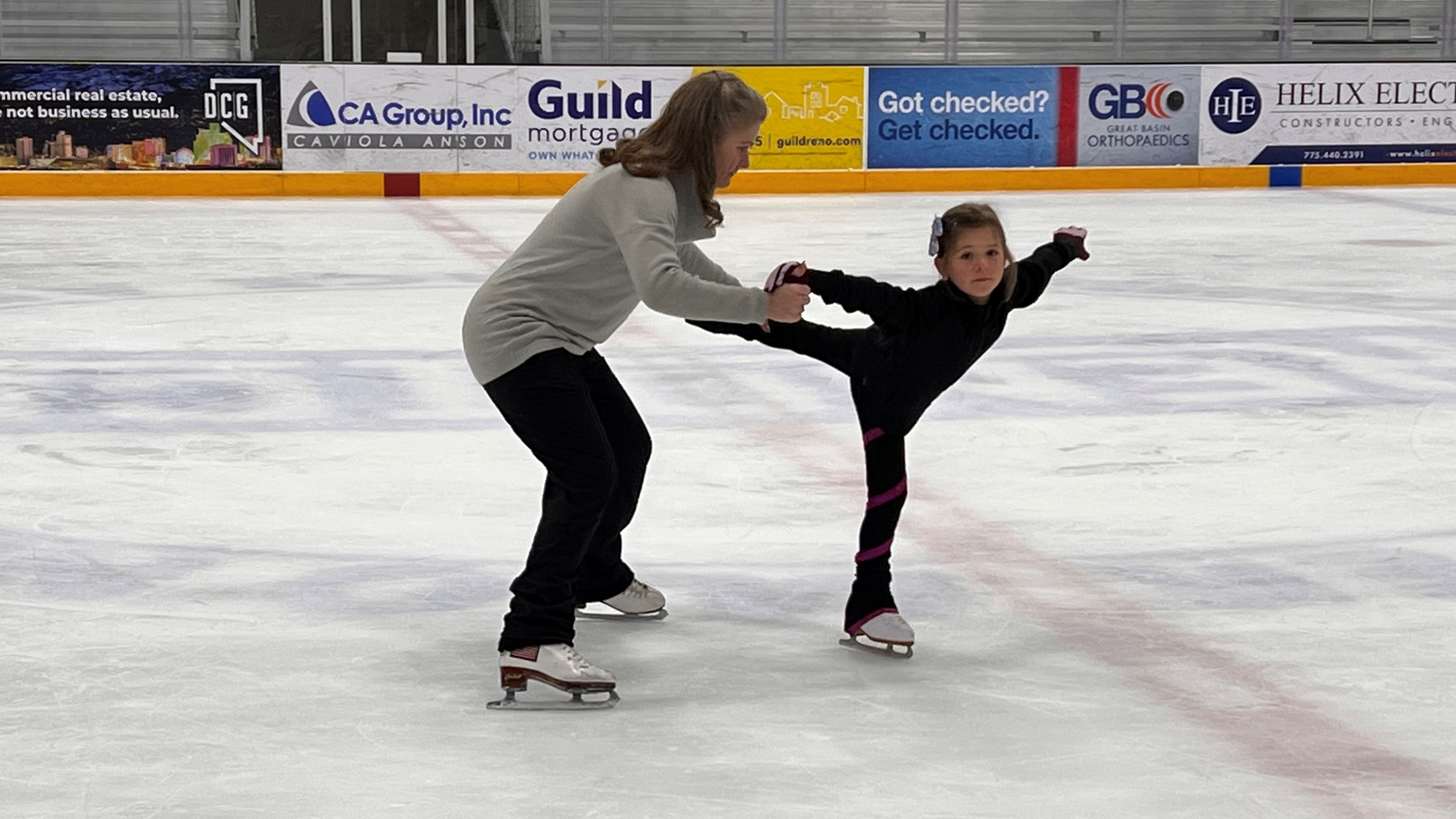 A coach assists a skater with a spiral. The coach is a white woman with shoulder length brown hair. She is wearing a grey long sleeve and black pants. The skater is a young white girl wearing a black long sleeve shirt and black pants with her blonde hair tied back in a ponytail