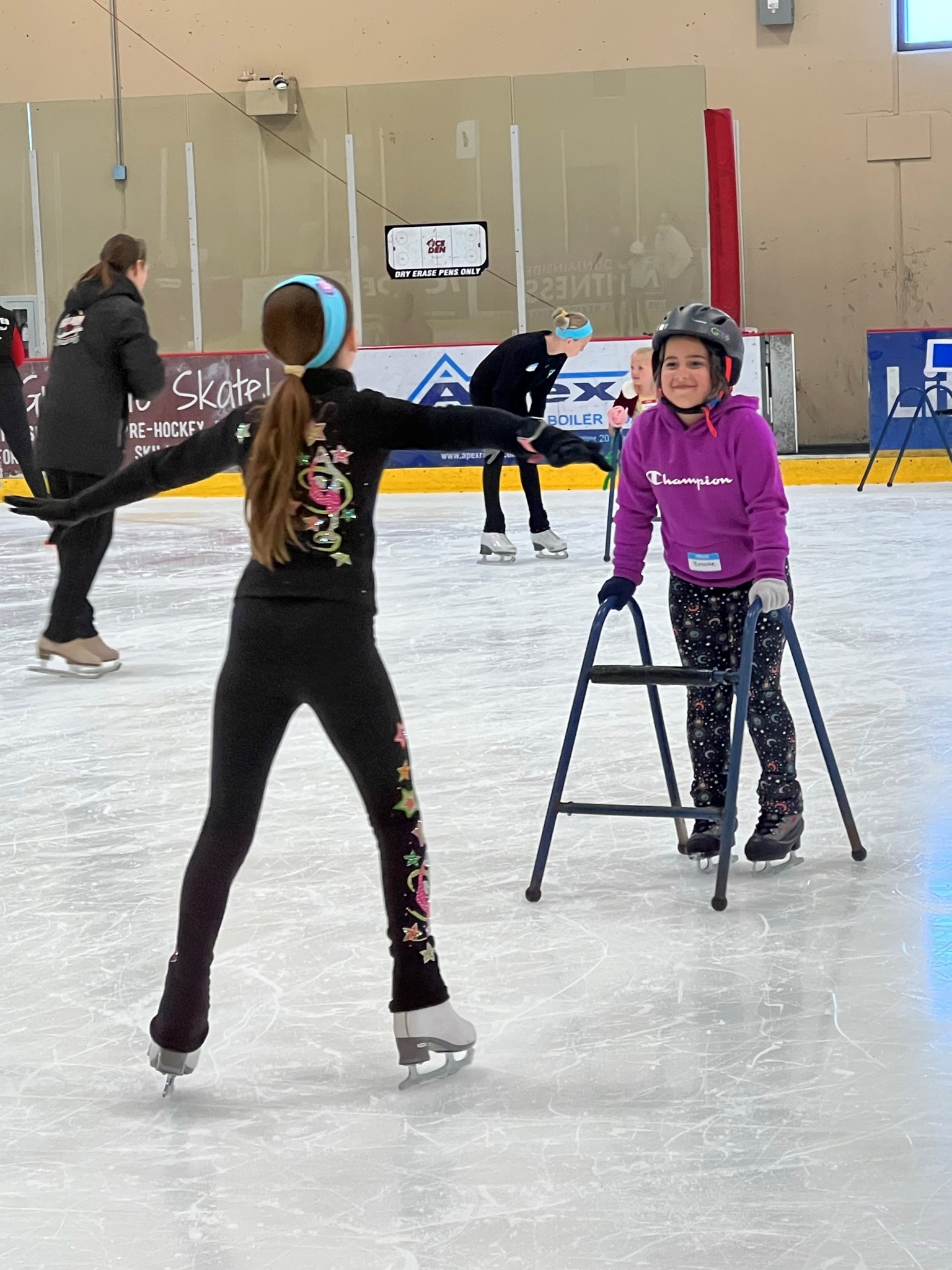 A volunteer helps a participant learn how to skate. The volunteer (left) has her back to the camera. She is wearing black pants with star details down the sides and a black jacket. She has long brown hair and is wearing a light blue headband. The skater (right) is wearing a black helmet, purple Champion hoodie and black pants with moons on them. She holds on to a walker to help stabilize herself on the ice