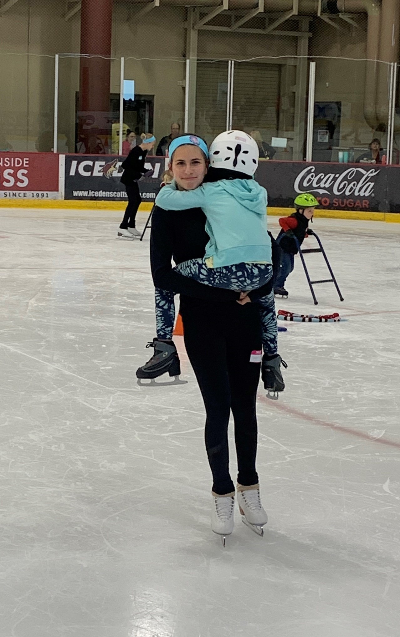 Sasha holds a participant of the event and smiles at the camera. Sasha is a young white woman with long brown hair tied back in a ponytail and a blue headband. She is wearing a black long sleeve shirt and black pants. The skater she is holding is wearing a white helmet, light blue jacket and monochrome blue tie dye pants
