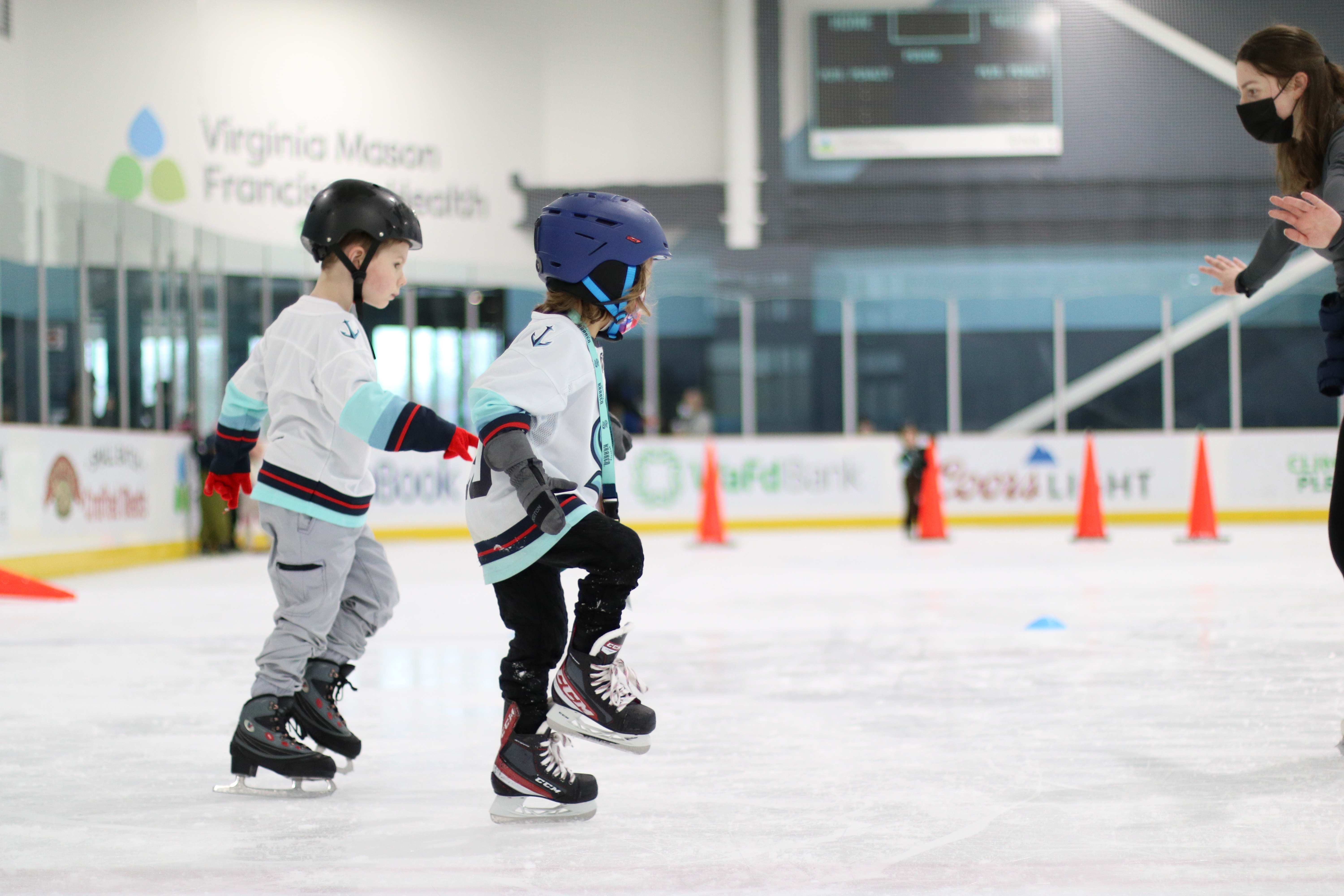 Two young skaters in hockey skates and a helmet hold up one leg while skating.