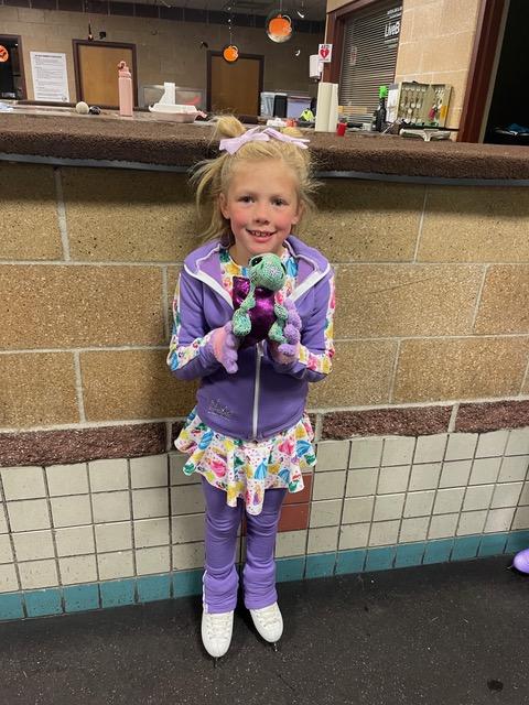 Photo of a young blonde girl wearing ice skated and smiling/ She is wearing a purple jacket and leggings with a multicolored skirt. She is holding a green and pink turtle stuffed animal.