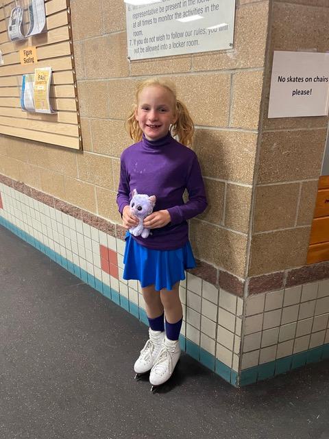 Photo of a young blonde girl wearing ice skates and smiling. She is wearing a purple long sleeve shirt and blue skirt.