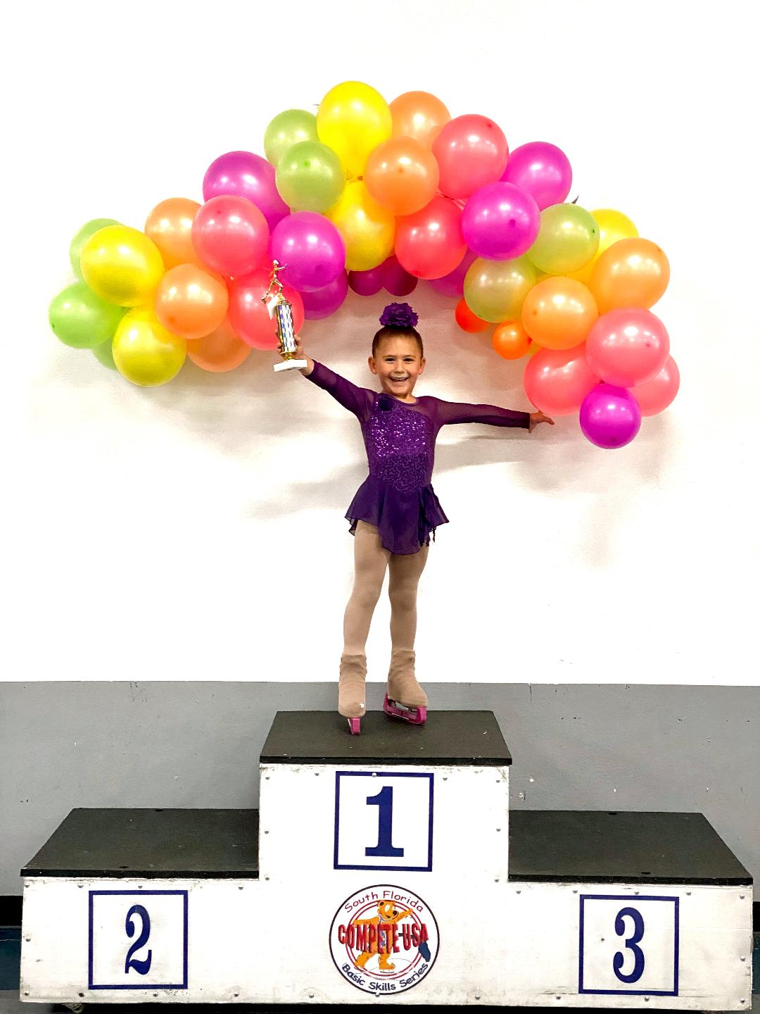 Photo of a young female skater standing on top of a podium and smiling. She is wearing a purple costume and has her hair in a tight bun. She is holding a trophy above her head in her right hand. Behind her is a balloon arch with green, yellow, orange and pink balloons