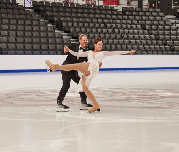 Donna and Kevin celebrate their first dance on ice. Donna has her brown hair tied up in a bun and she is wearing a white long sleeve skating costume. She kicks one leg out in the air and extends one arm outward. Kevin stands behind her holding one hand on her waist. Kevin is wearing a black suit with a black bowtie and hockey skatess