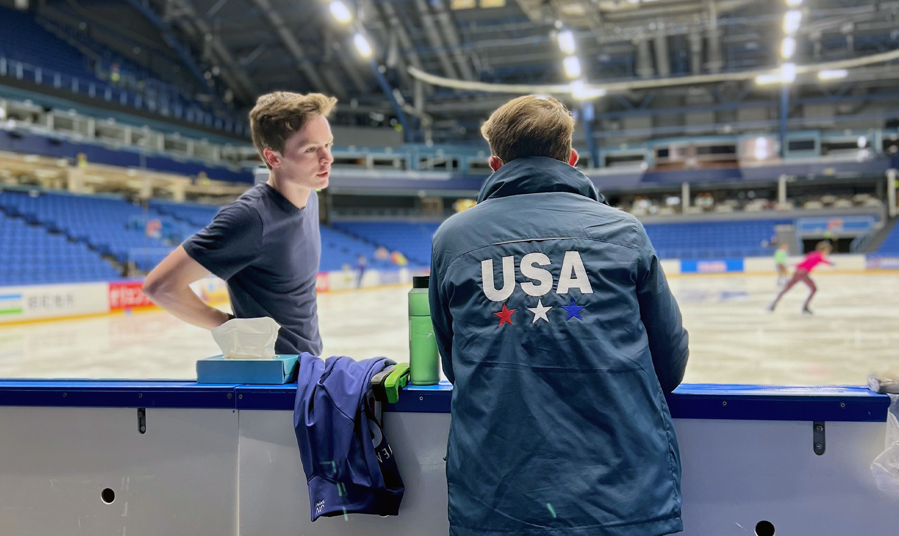 A coach stands with his back turned to the camera, instructing an athlete on the ice from behind the boards.