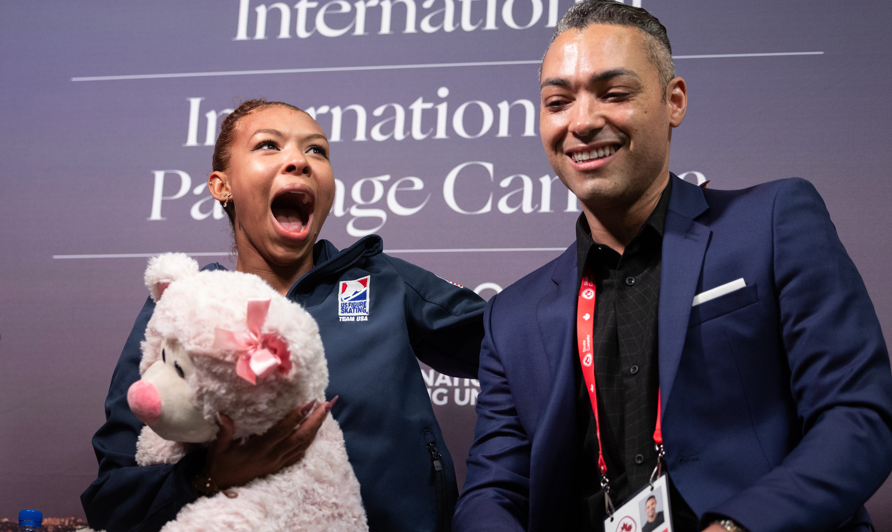 Starr Andrews gives a stunned reaction to her score in the kiss and cry while she holds a large white teddy bear. She is wearing a blue Team USA jacket
