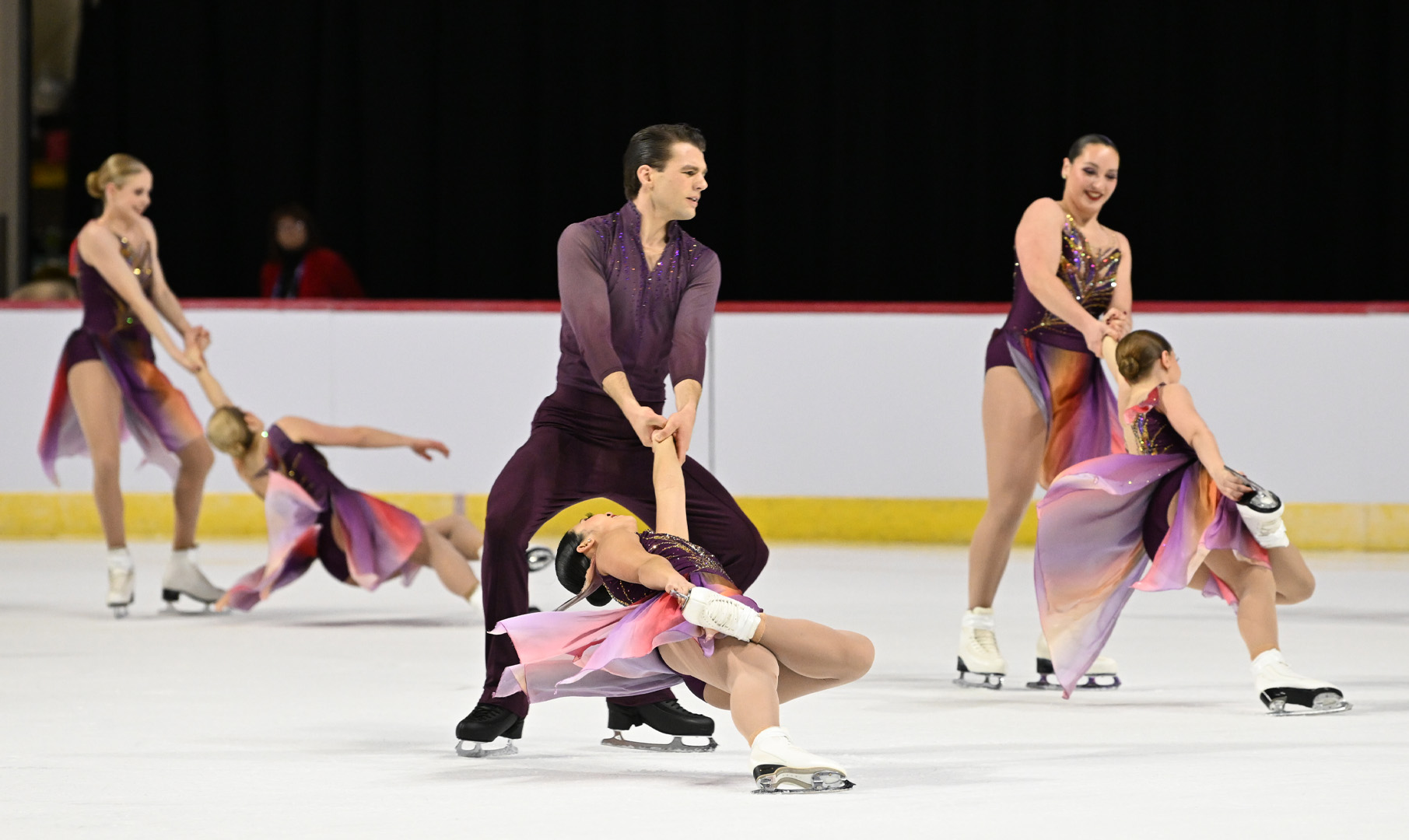 The Haydenettes perform a pair element wearing matching purple costumes. In the foreground of the photo is a man wearing a 3/4 sleeve purple top and black pants, holding on to his teammate who is a woman wearing a matching purple dress.