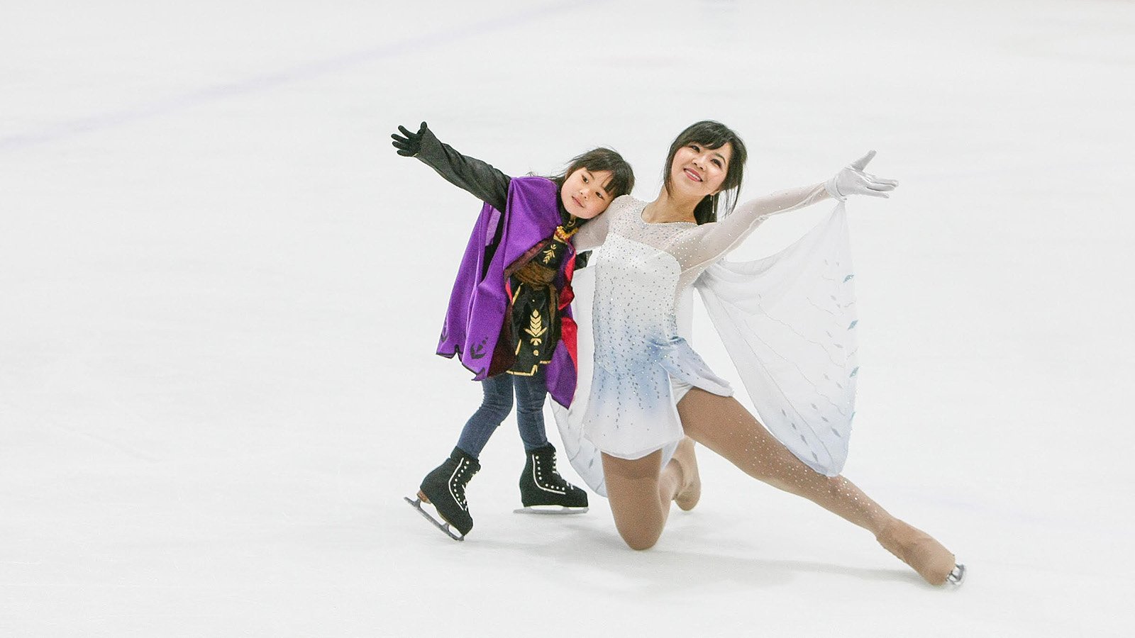 Mother Ashley Chen, on one knee wearing a white dress, embraces her daughter, wearing a black costume with a purple top, while skating together on the ice.