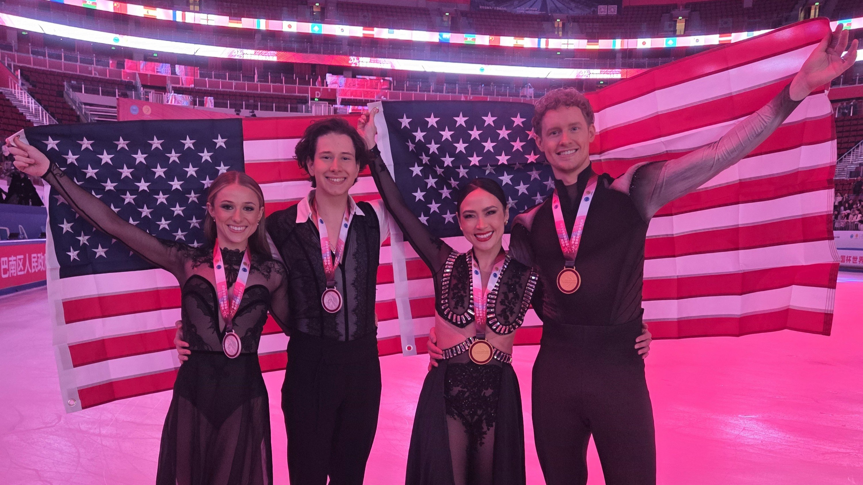 From left to right: Emilea Zingas, Vadym Kolesnik, Madison Chock, and Evan Bates stand smiling at the camera, holding two American flags behind them with medals from 2025 Cup of China around their neck.