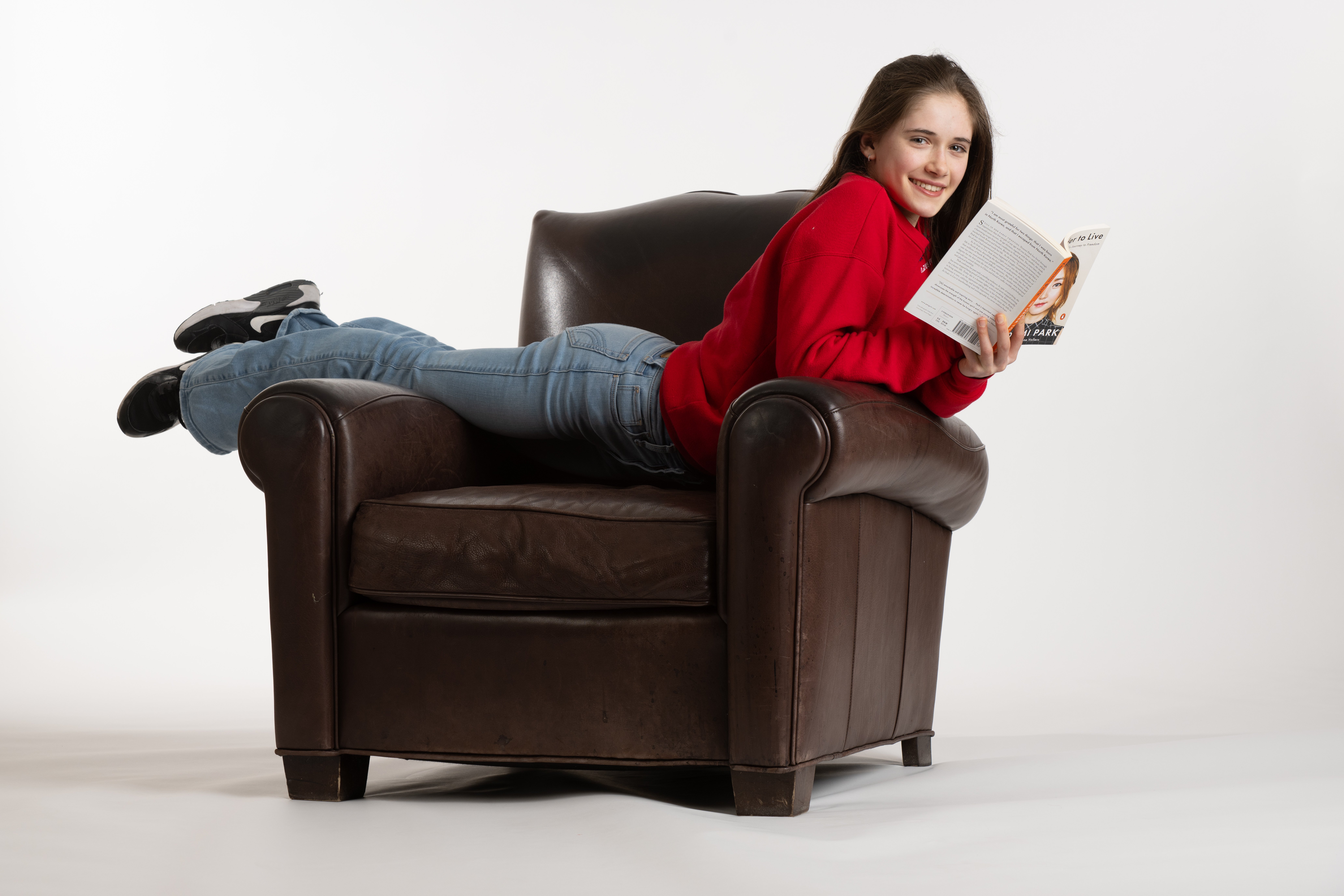 Isabeau Levito poses laying across a brown leather chair holding a book. She is a young woman with long brown hair wearing a red long sleeve shirt and light blue jeans