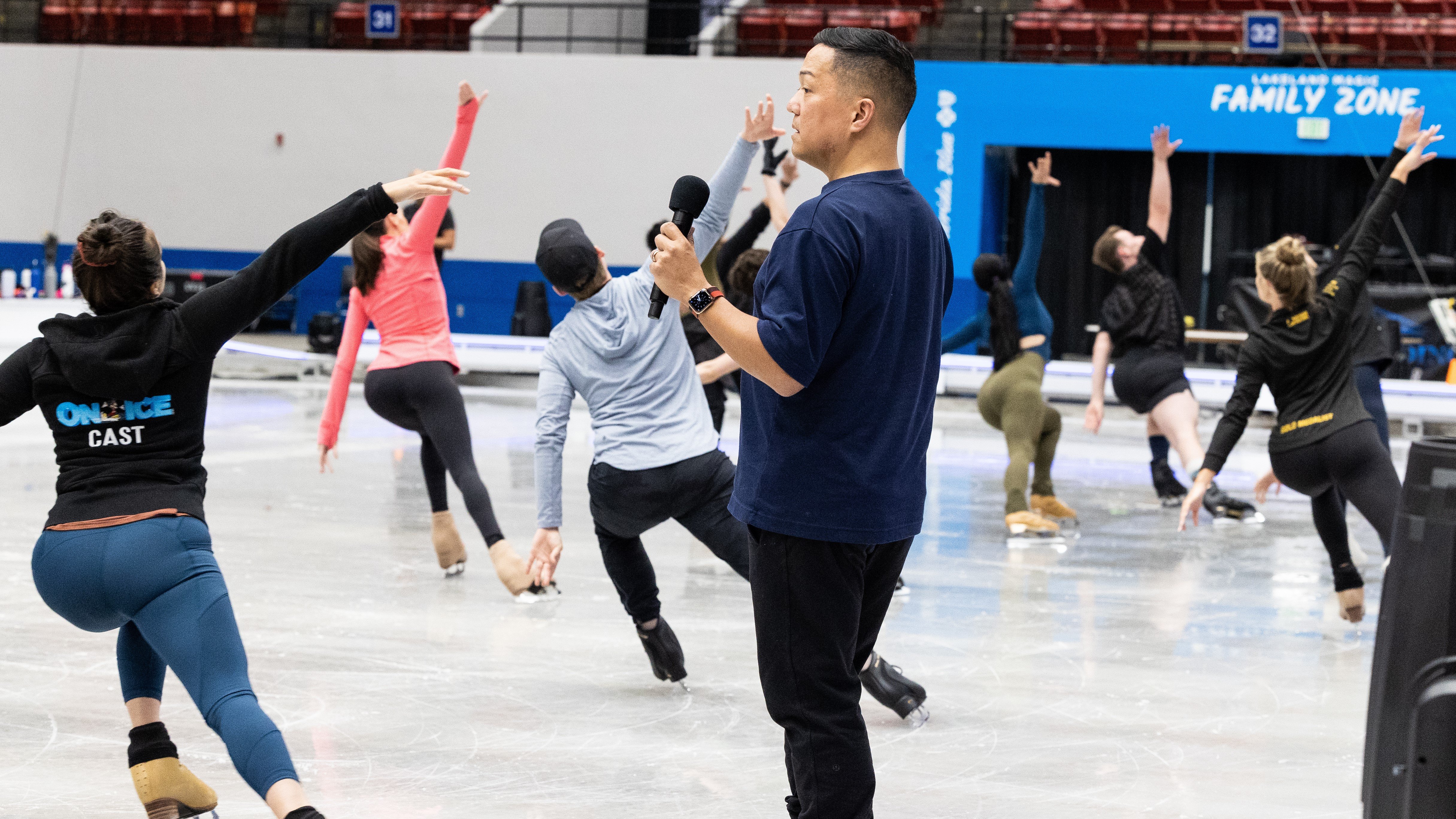 Gig stands in the forefront of the photo with a mic in his hand, facing skaters who are practicing Disney on Ice  choreography.