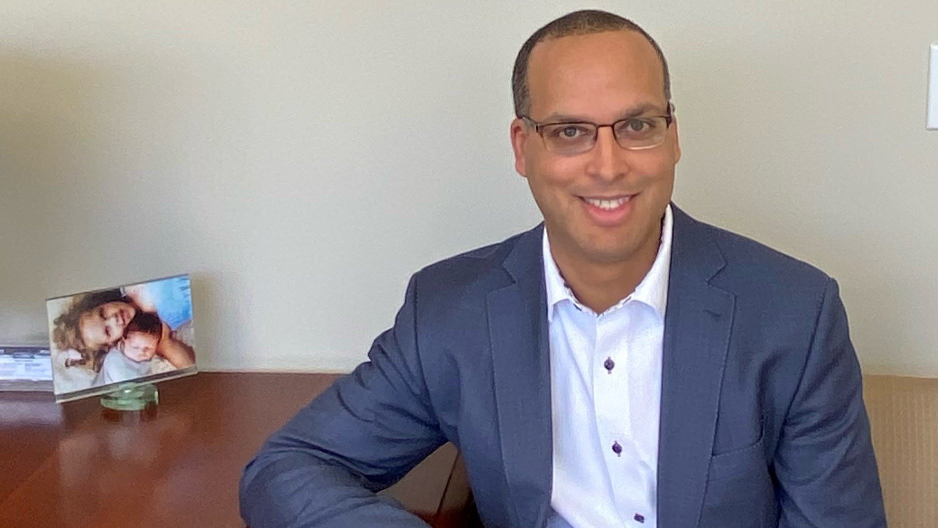Aaron Parchem is an African American man wearing a blue suit with a white collared shirt underneath and glasses. He sits at a desk with his right elbow on the table. A photo of his young kids sits on the desk behind him