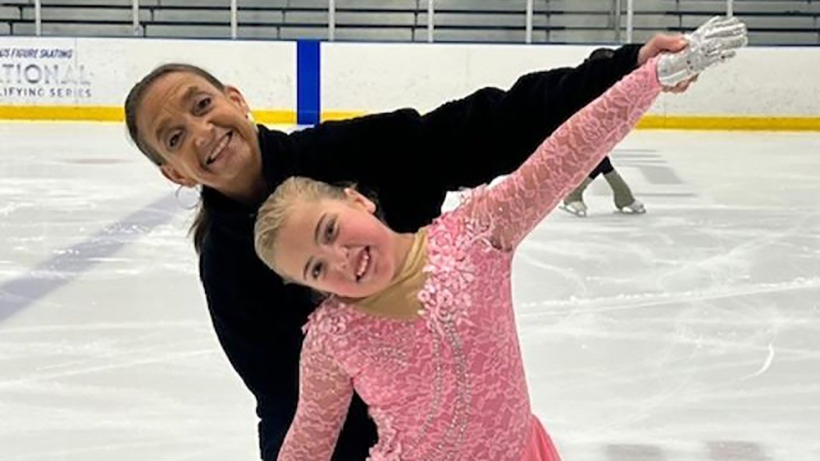 Coach Jane Fransen, dressed in all black, stands behind Blakely Schroeder, dressed in pink, embraced with arms extended.