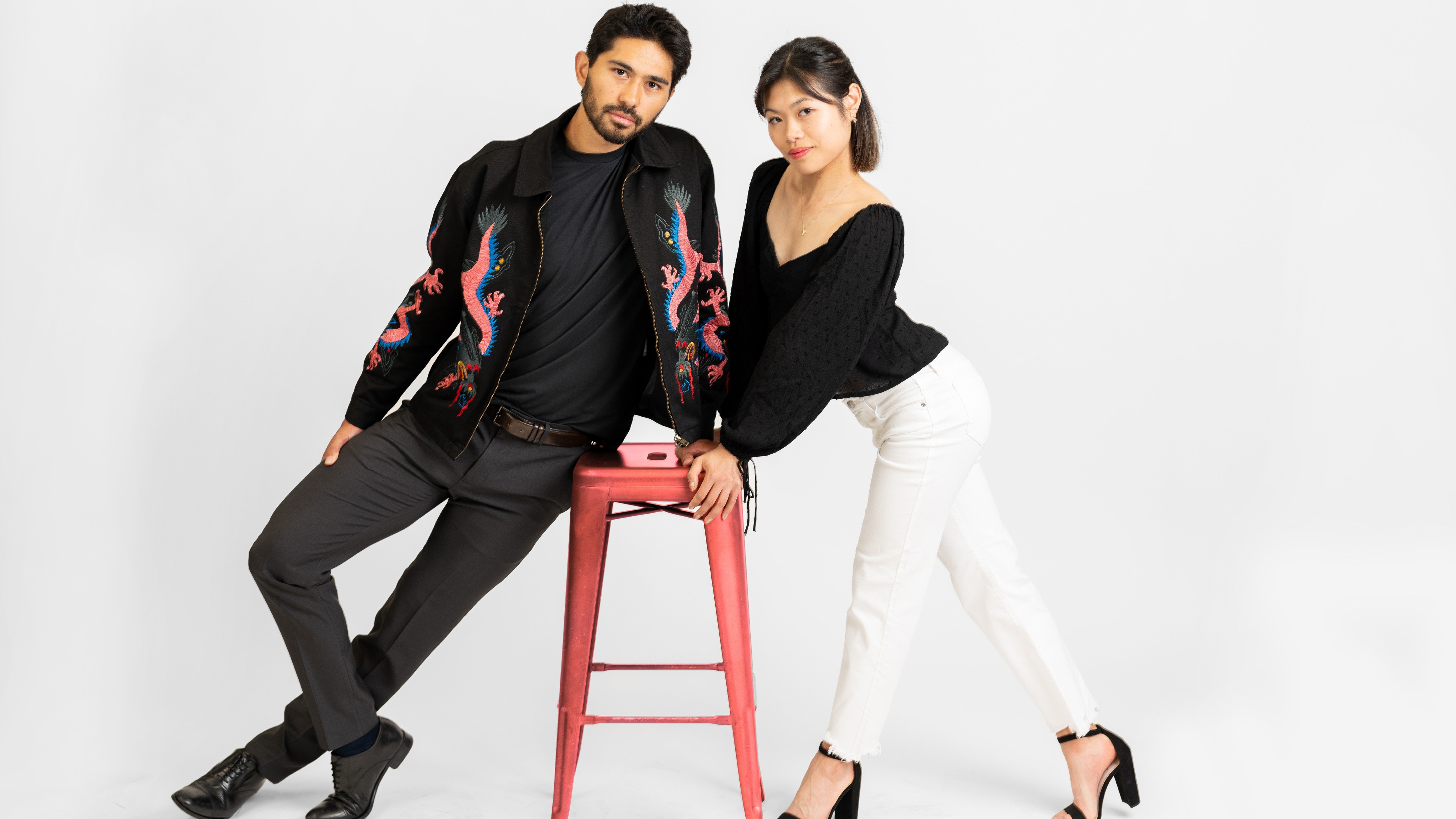 Emily Chan and Spencer Howe learn on a stool while posing for their cover shoot for SKATING magazine. Howe (left) is a man with short black hair wearing a black jacket with a bright pink and blue dragon design over a black shirt and black jeans. Chan is a woman with short black hair wearing a black long sleeve shirt, white jeans, and black high heels 
