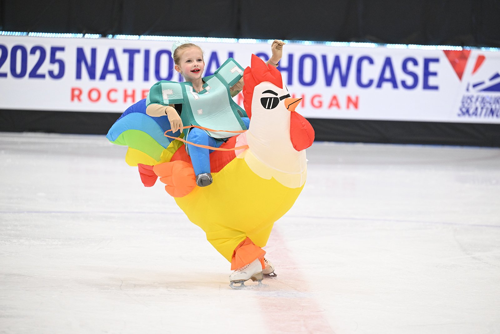 Katarina Suemnig rides an inflatable yellow chicken. The 2025 National Showcase banner is in the background. She's wearing a green costume with padding. 