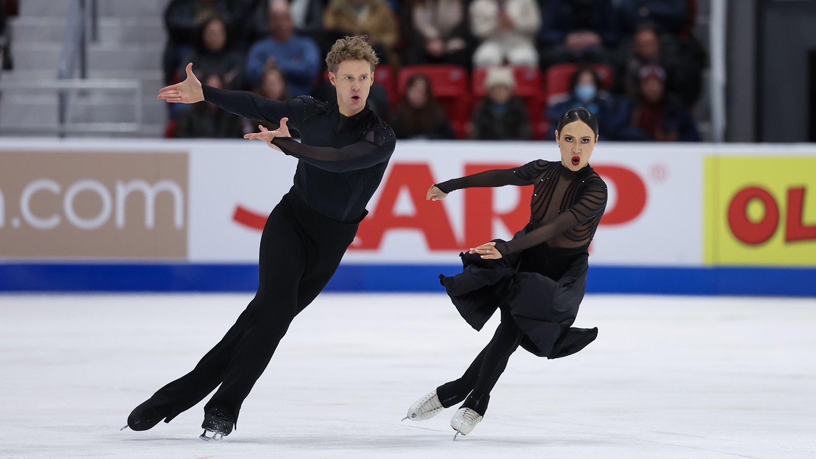 Madison Chock and Evan Bates skate side by side with both athletes pointing their arms to the left in unison. Evan (left) is a tall man with short blonde hair wearing a long sleeve black top and black pants. Madison (right) is a woman with black hair tied back in a bun. She is wearing a black skating costume with long sleeves and a long black skirt