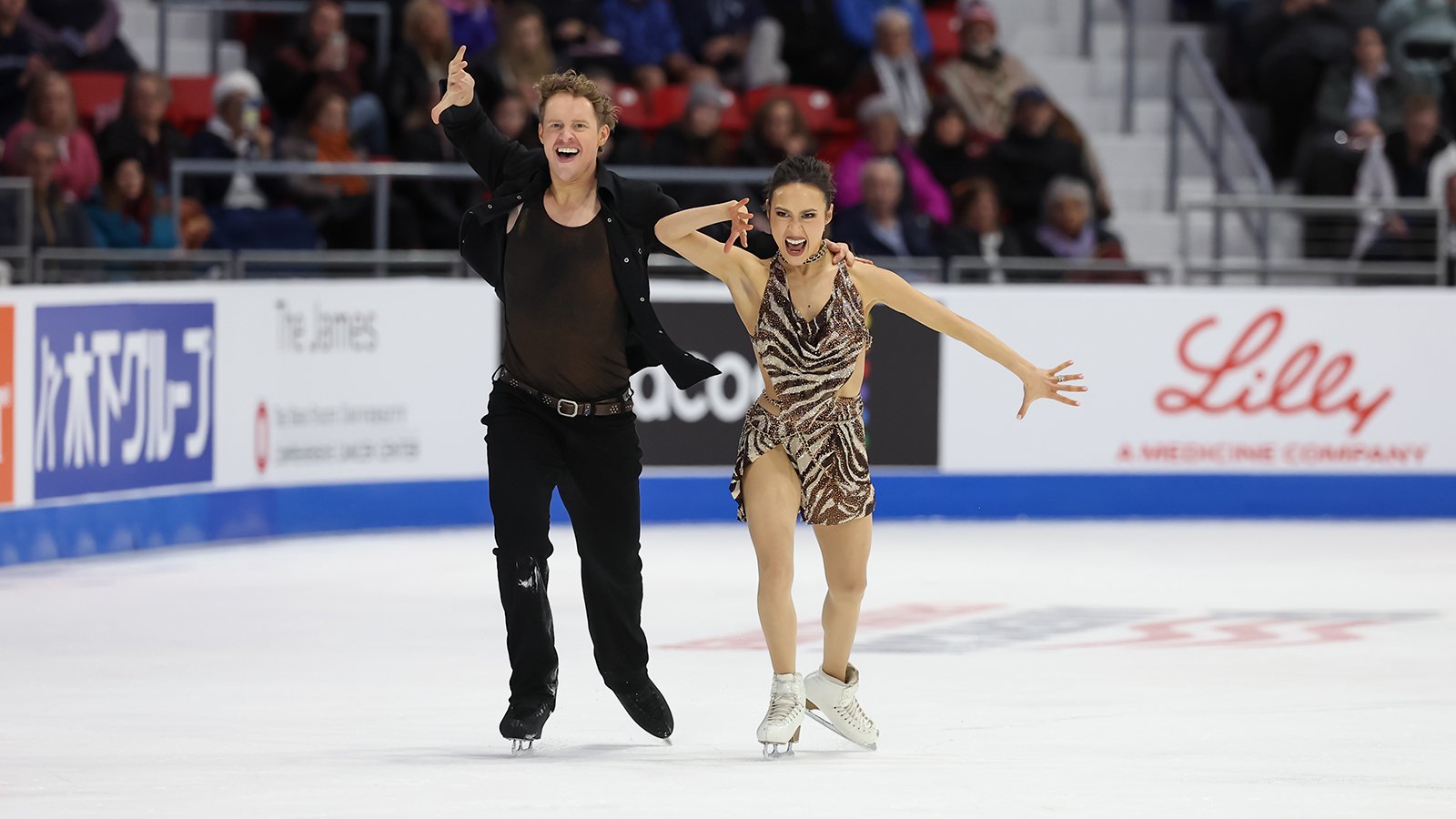 Madison Chock and Evan Bates perform their rhythm dance at 2025 Saatva Skate America. They skate side by side with their right hands raised to their face in unison with a determined look on their face. Evan (left) is a tall man with short blonde hair wearing a brown tank top with a black button down shirt over top and black pants. Madi is a woman with long black hair wearing a matching tiger print top and skirt