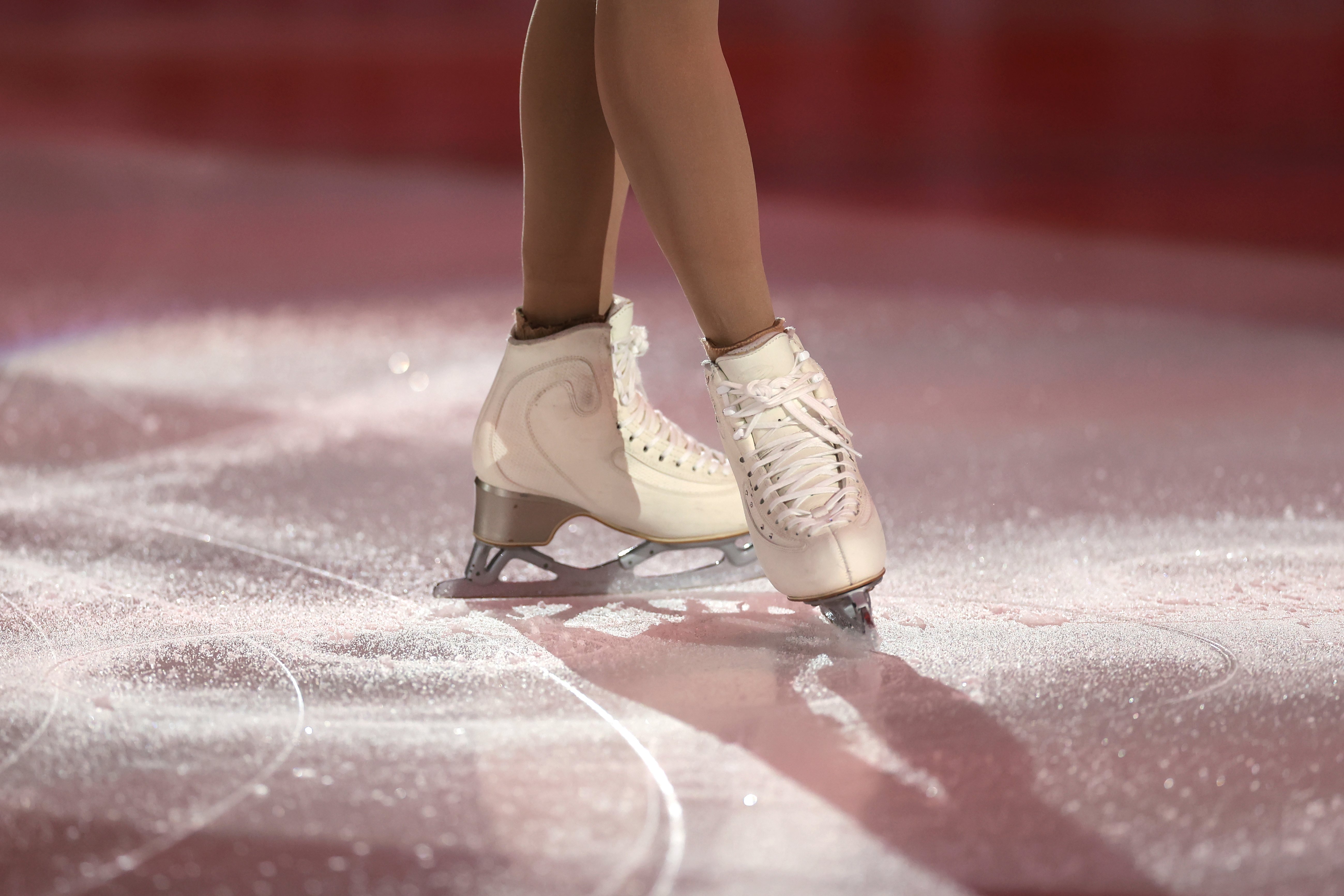 Photo of a skaters legs on the ice wearing white skates. A red light shines over the ice. 