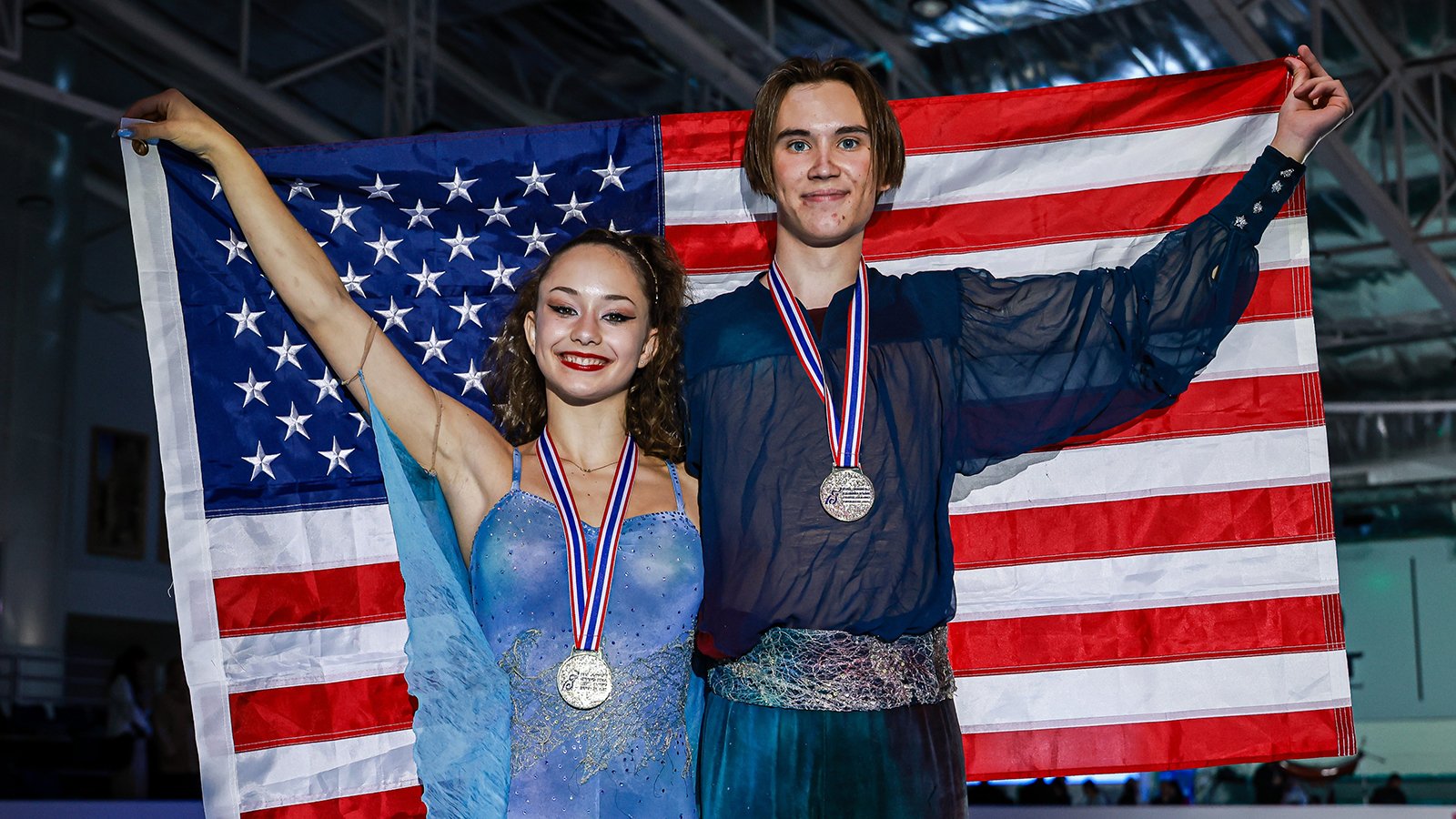 Hana Maria Aboian, wearing blue, and Daniil Veselukhin, wearing black, hoist the American flag after receiving their gold medals at JGP Thailand.