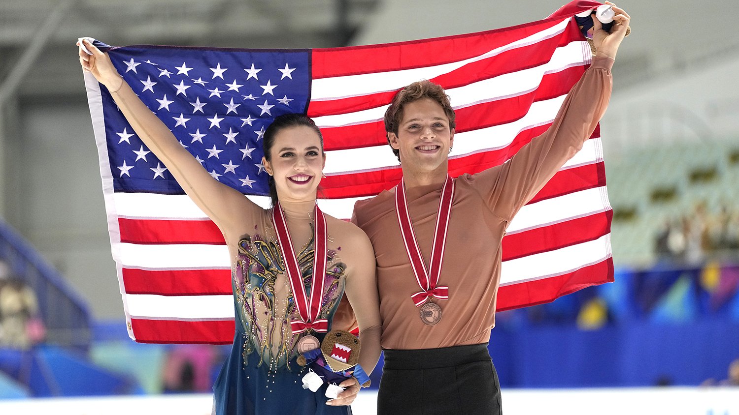 Caroline Green (left) and Michael Parsons (right) hold up the U.S. flag behind them and smile during the medal ceremony at NHK Trophy. They have bronze medals around their neck and they both smile.