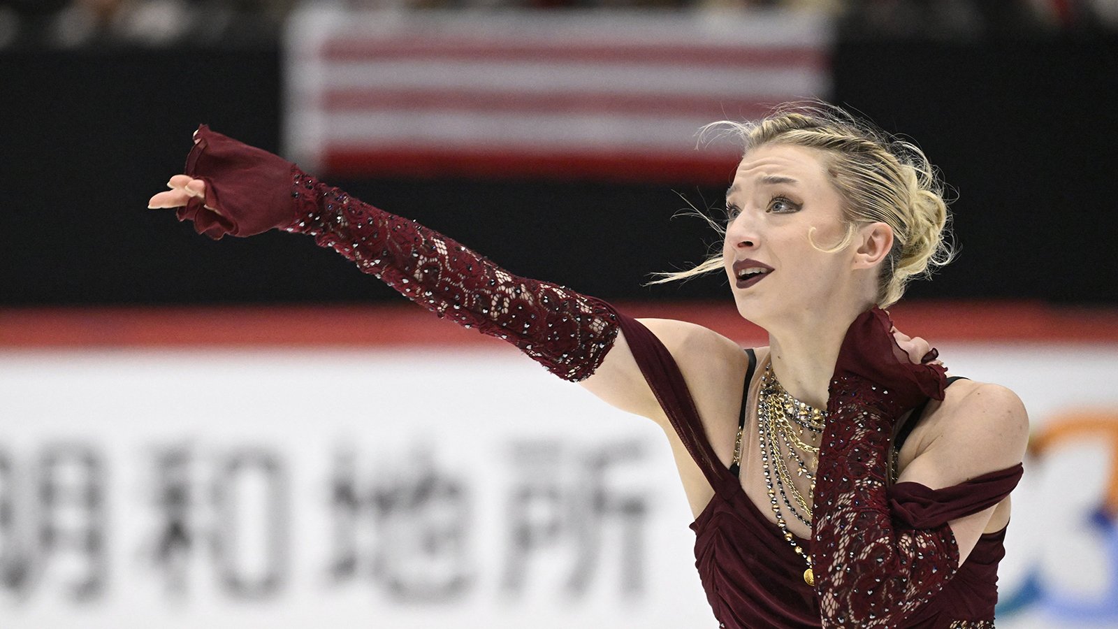 Photo taken from the waist up of Amber Glenn. She looks off to the left with one arm outstretched. Amber is a young woman with blonde hair tied back in a bun wearing a maroon skating costume with chunky gold necklaces and matching maroon gloves.