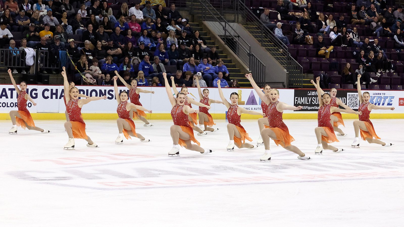 Photo of the Ice Denettes Affinity intermediate team. All athletes are in a coordinated kneeling position with their arms extended and smiles on their faces. They are all wearing matching skating costumes with a red jeweled top and orange skirt. 