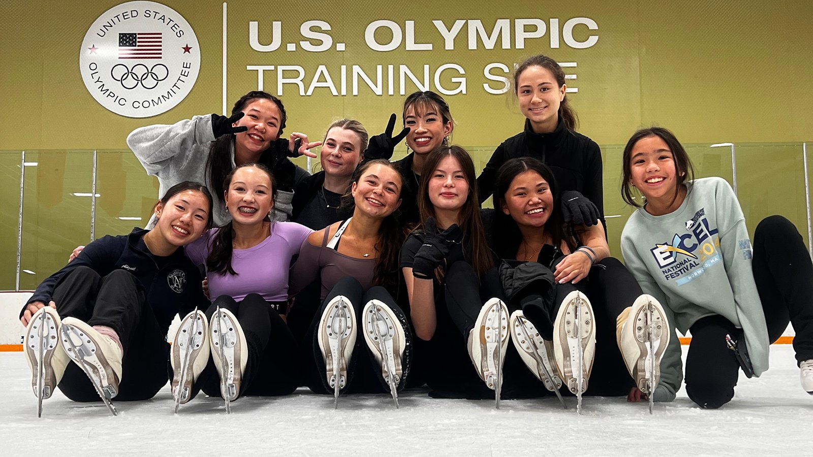 A group of female skaters pose for a photo on the ice on front of a sign that says "U.S. Olympic Training Site"