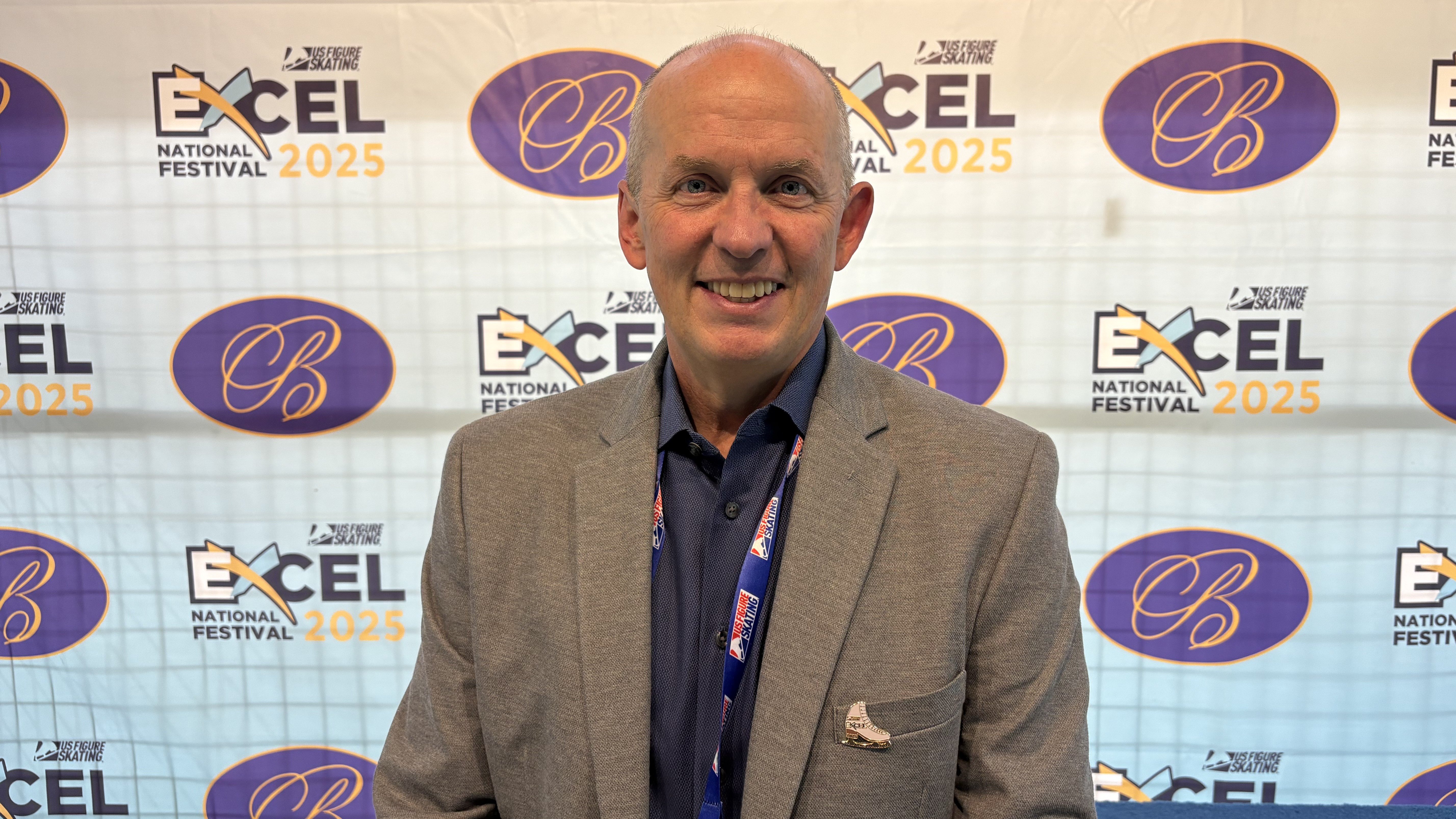 Photo of U.S. Figure Skating CEO Matt Farrell. Matt is an older white man wearing a navy blue button down shirt and a grey blazer. He stands in front of a backdrop with the 2025 Excel National Festival logo and the Broadmoor logo on it