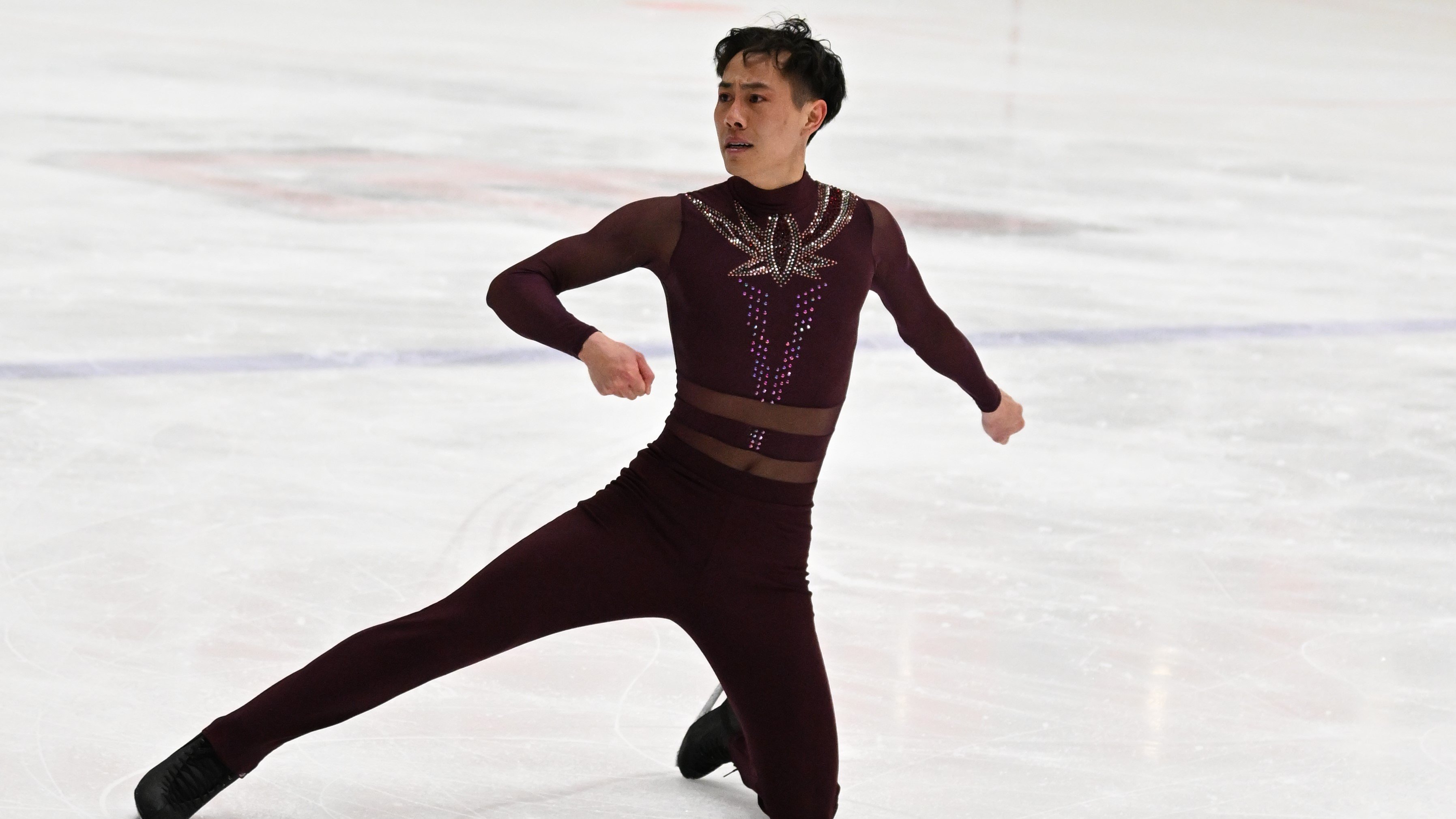 JY competes on the ice. They are an Asian skater with short hair wearing a maroon skating costume with jewels down the front. They are kneeling on their left knee with their right leg outstretched to the side
