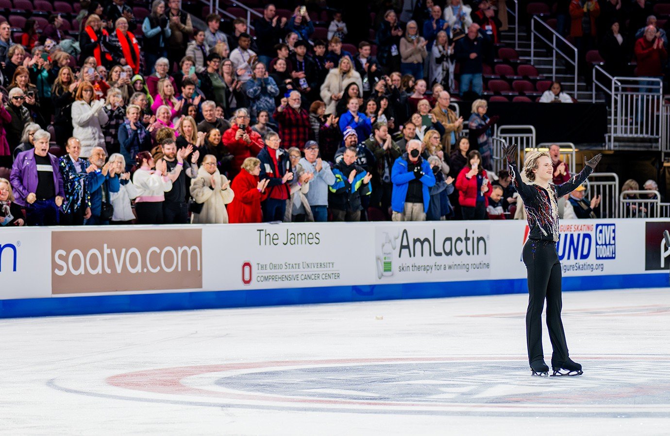 Ilia Malinin salutes the crowd with his arms outstretched and a smile on his face as people in the stands cheer. Ilia is a young man with short blonde hair wearing a black top with red and white stripes and black pants. 