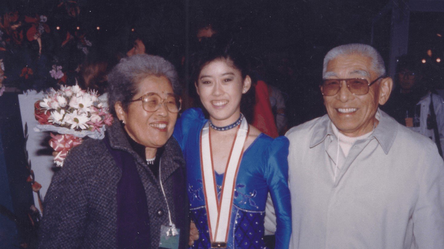 Kristi poses for a photo with her grandparents. Kristi (center) is an Asian woman wearing a blue skating costume with puffy sleeves and a medal around her neck. Her grandmother (left) is an older Asian woman with short grey hair and glasses. She is wearing a grey coat. Her grandfather (right) is an older Asian man with grey hair and glasses. He is wearing a white button up shirt 