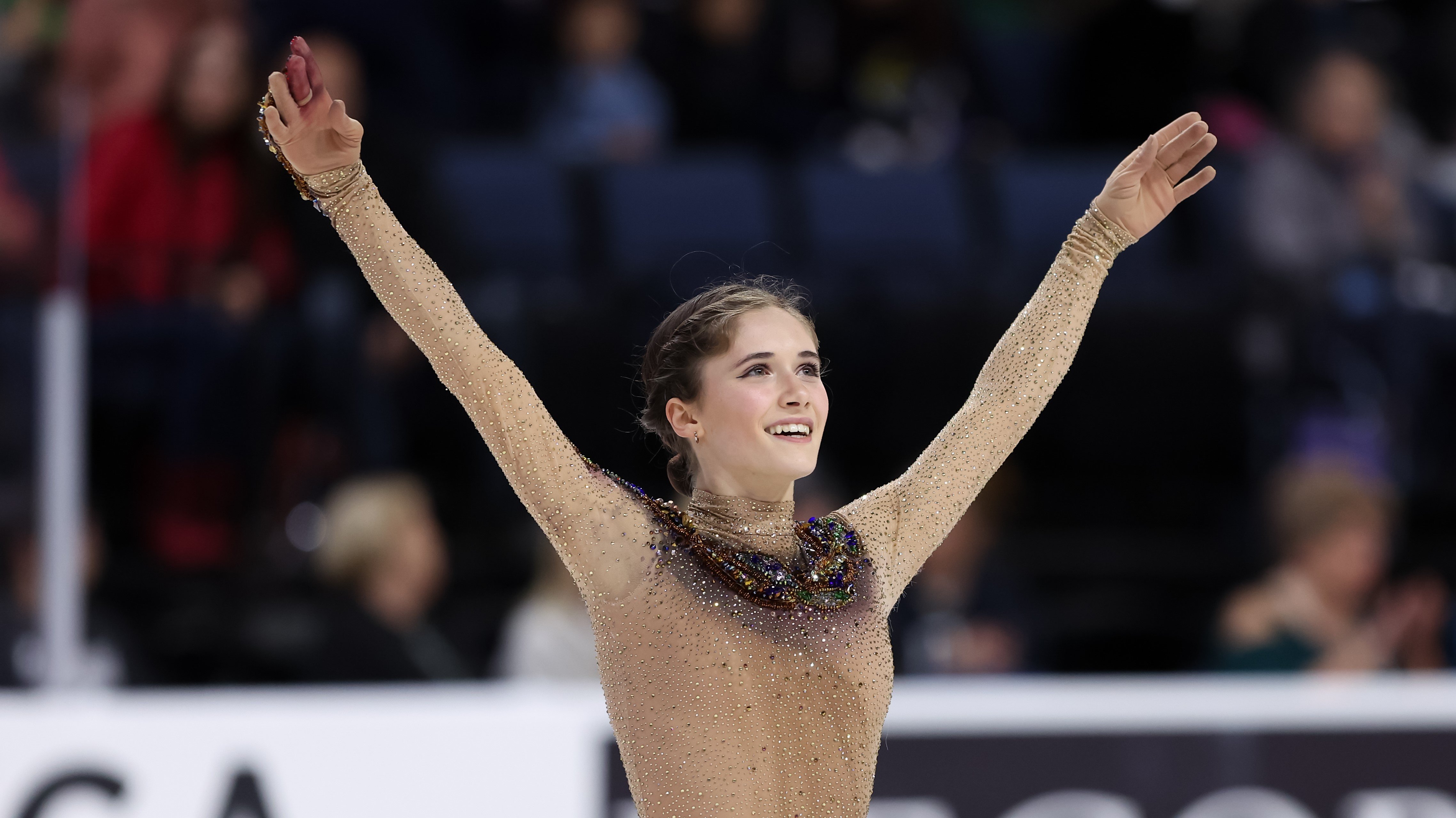Isabeau Levito, wearing a light brown colored dress with a snake on it, raises her arms as she salutes the crowd.