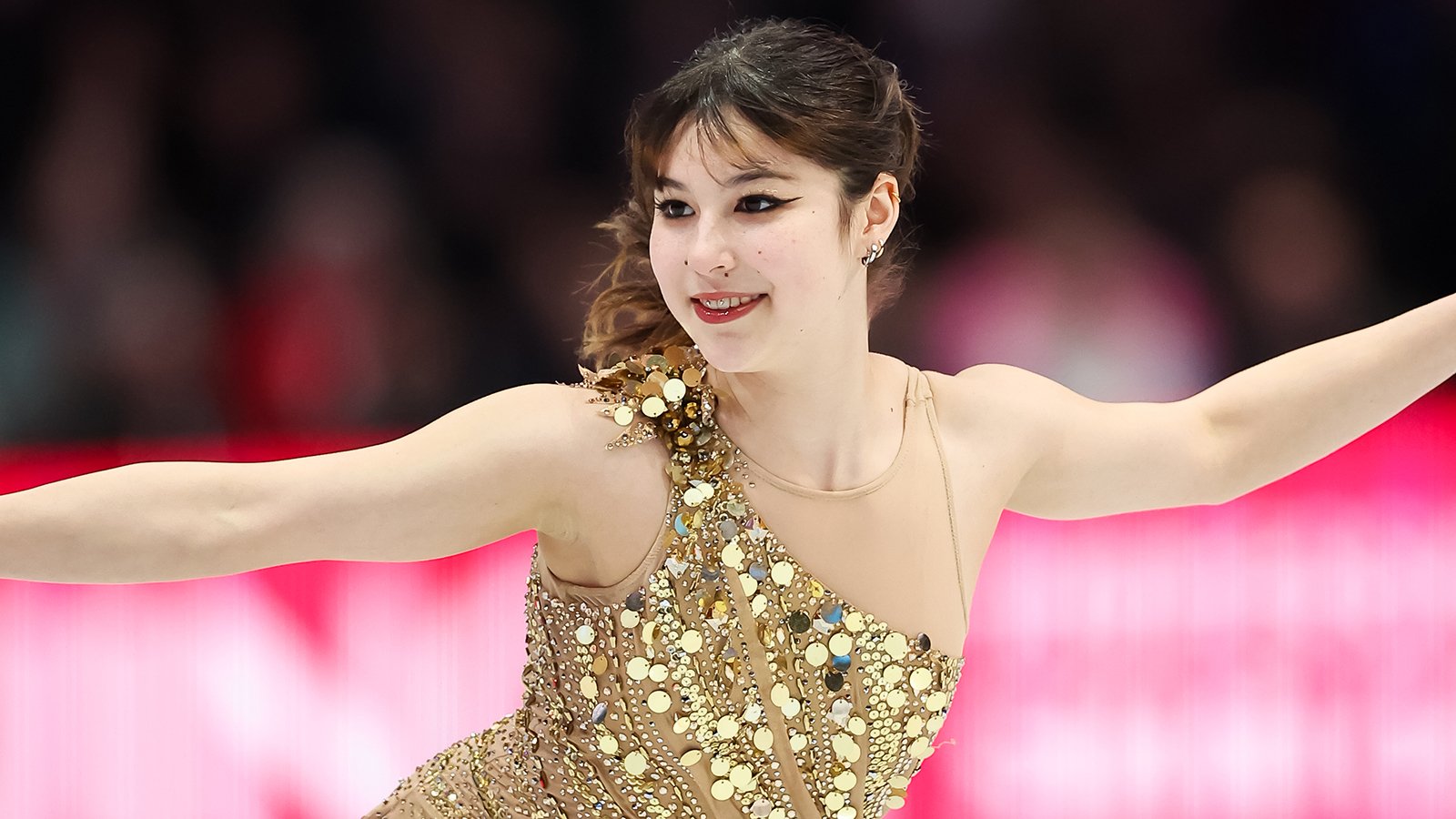 Photo of Alysa Liu taken from the waist up. She is a young woman with brown hair tied back in a ponytail wearing a gold sparkly dress. She stands with her arms outstretched, looking to the left with a slight smile on her face. 