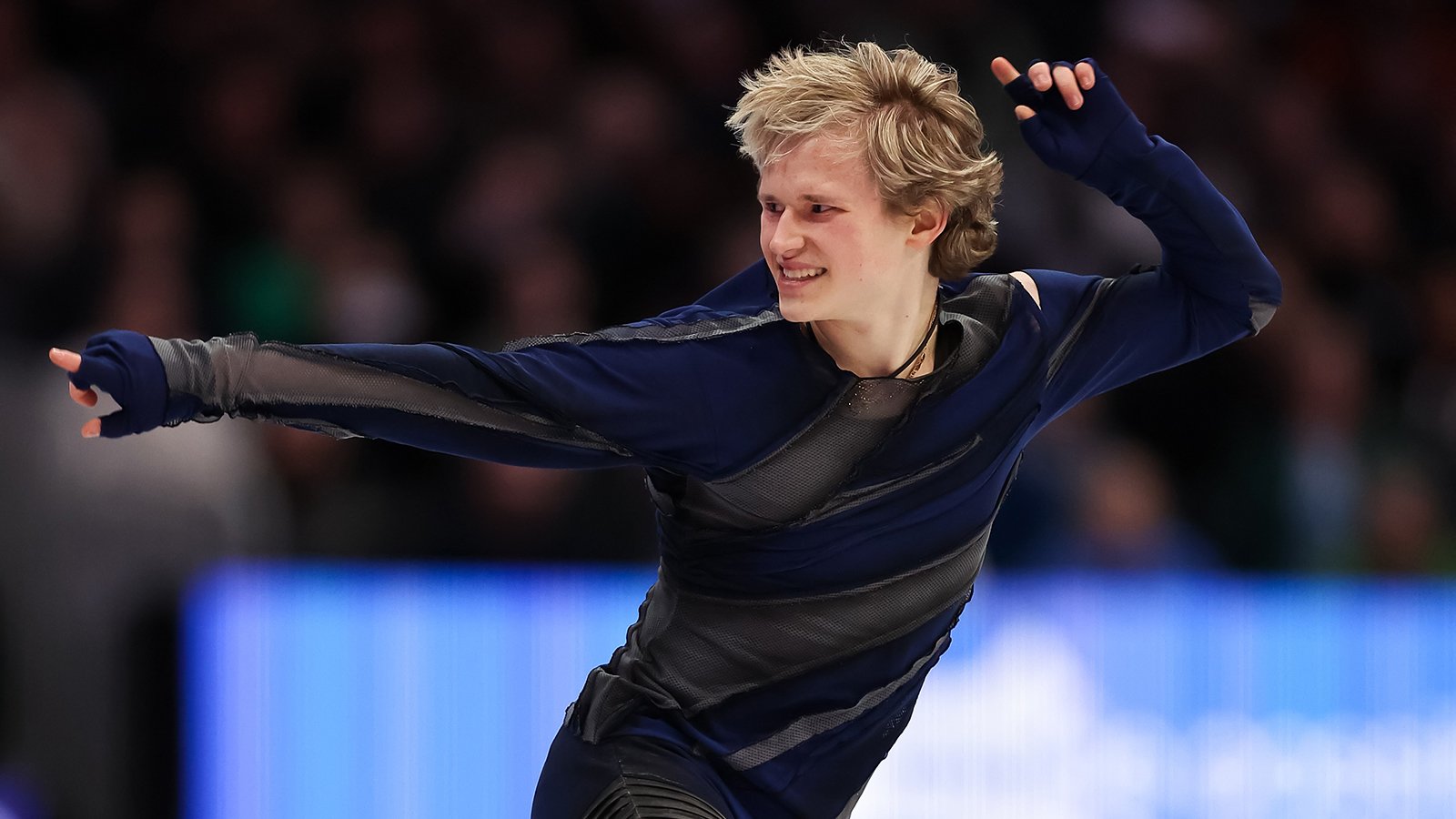 Dressed in all black, Ilia Malinin performs a routine. He is smiling and pointing straight out with one arm and has the other arm pointing up