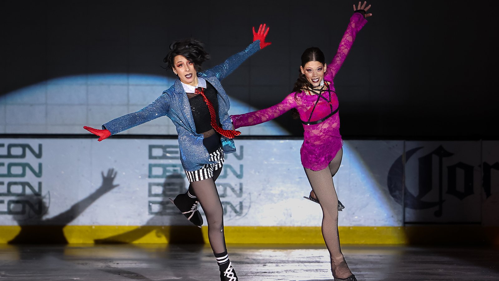 Ashlee Pantano (left) and Ashlyn Olson (right) skate side by side with their arms extended outward. Ashlee is wearing a black collared shirt with a red tie and a sparkly blue blazer over top with black and white striped shorts. Ashlyn is wearing a hot pink top with a black strap over her chest. 