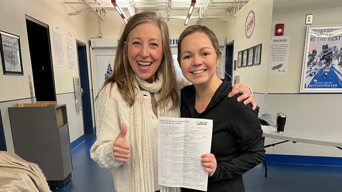 Sandy Lenz (left) poses for a photo from the waist up with her adult student, Emily Thompspon (right). Lenz is an older white woman with shoulder length blonde hair. She is wearing a white sweater with a white scarf and gives the camera a thumbs up with her other arm around the shoulder of her student. Emily is wearing a black long sleeve skating costume and it proudly holding up her testing papers with a big smile. 